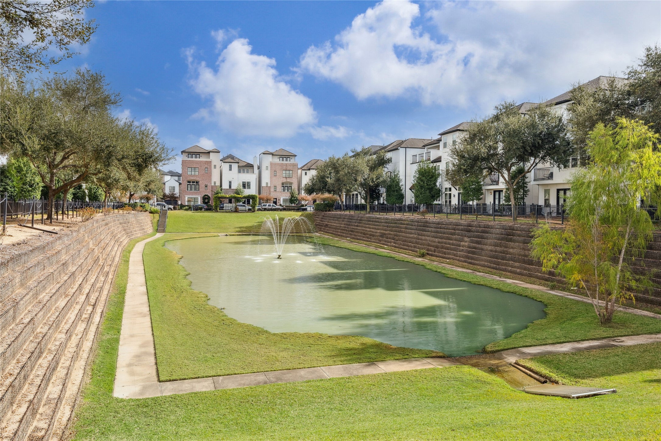 Landscaped community greenbelt centered around a tranquil pond with a working fountain, walking paths, and terraced stone walls. Surrounded by mature trees and well-kept residences, this shared outdoor space adds a peaceful, park-like setting to the neighborhood.