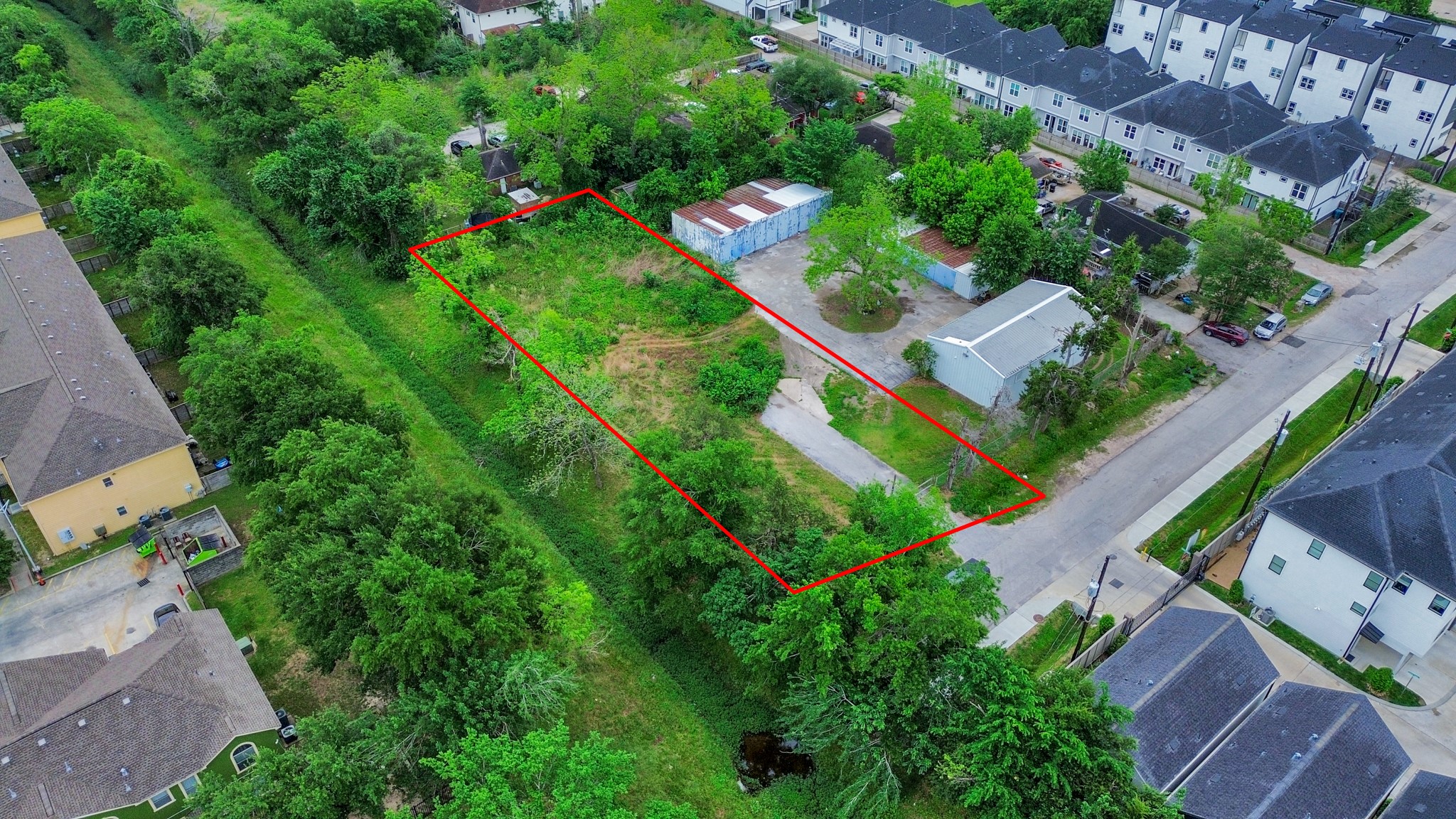 This aerial photo shows a rectangular plot of land outlined in red, surrounded by greenery and residential buildings. The lot appears to have a driveway and some structures, offering potential for development in a quiet neighborhood setting. This aerial photo shows a rectangular plot of land outlined in red, surrounded by greenery and residential buildings. The lot appears to have a driveway and some structures, offering potential for development in a quiet neighborhood setting.