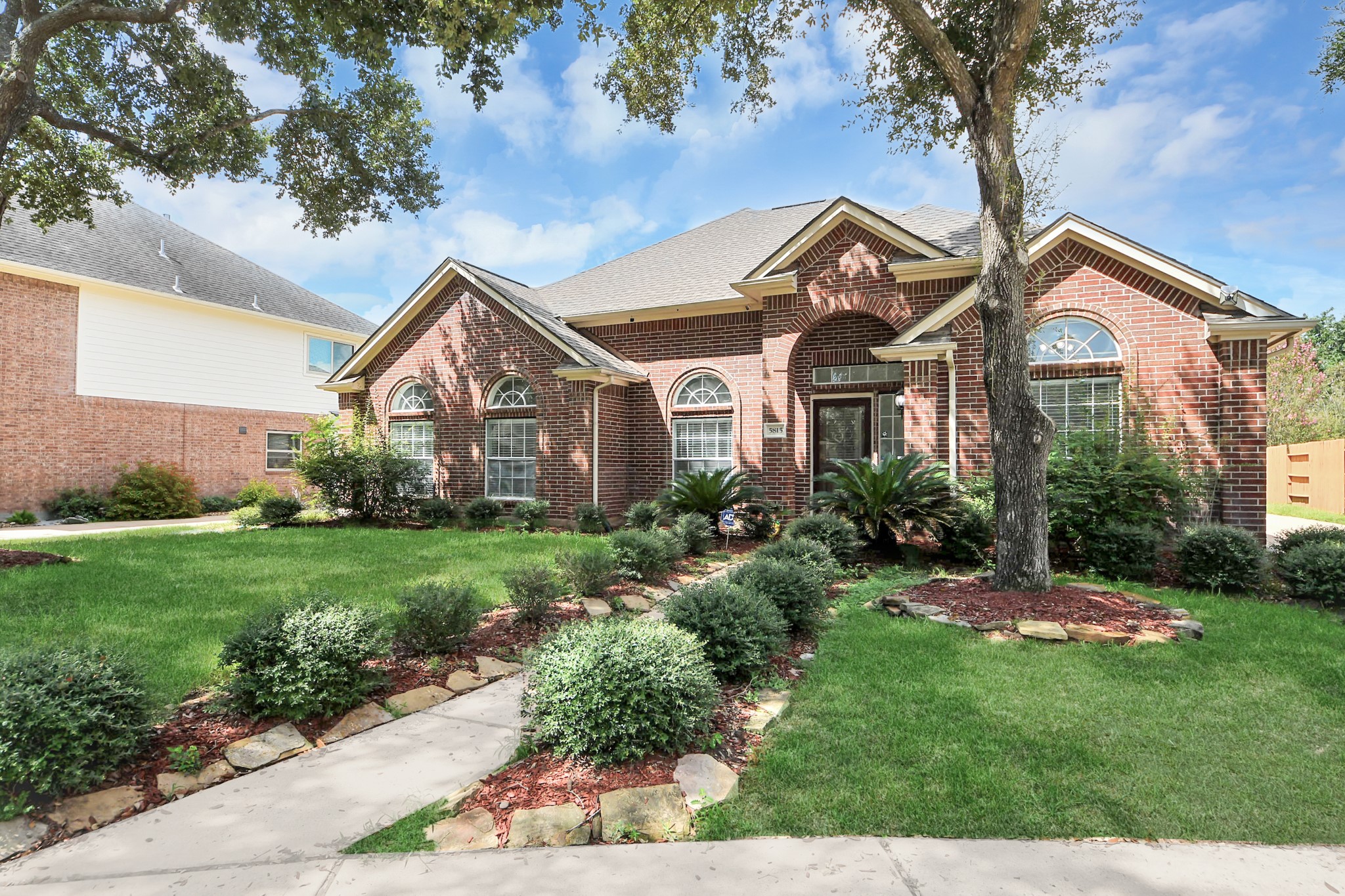 Charming brick home with beautiful arched windows, nestled in a lush landscaped garden featuring mature trees and manicured shrubs for added curb appeal.