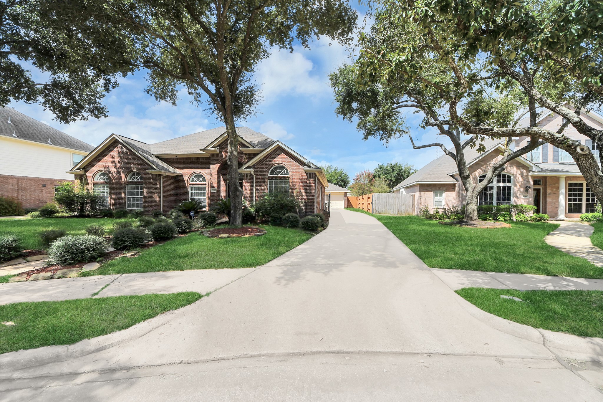 Charming brick home with elegant arched windows, nestled among mature trees and lush landscaping, featuring a wide driveway leading to a detached garage.