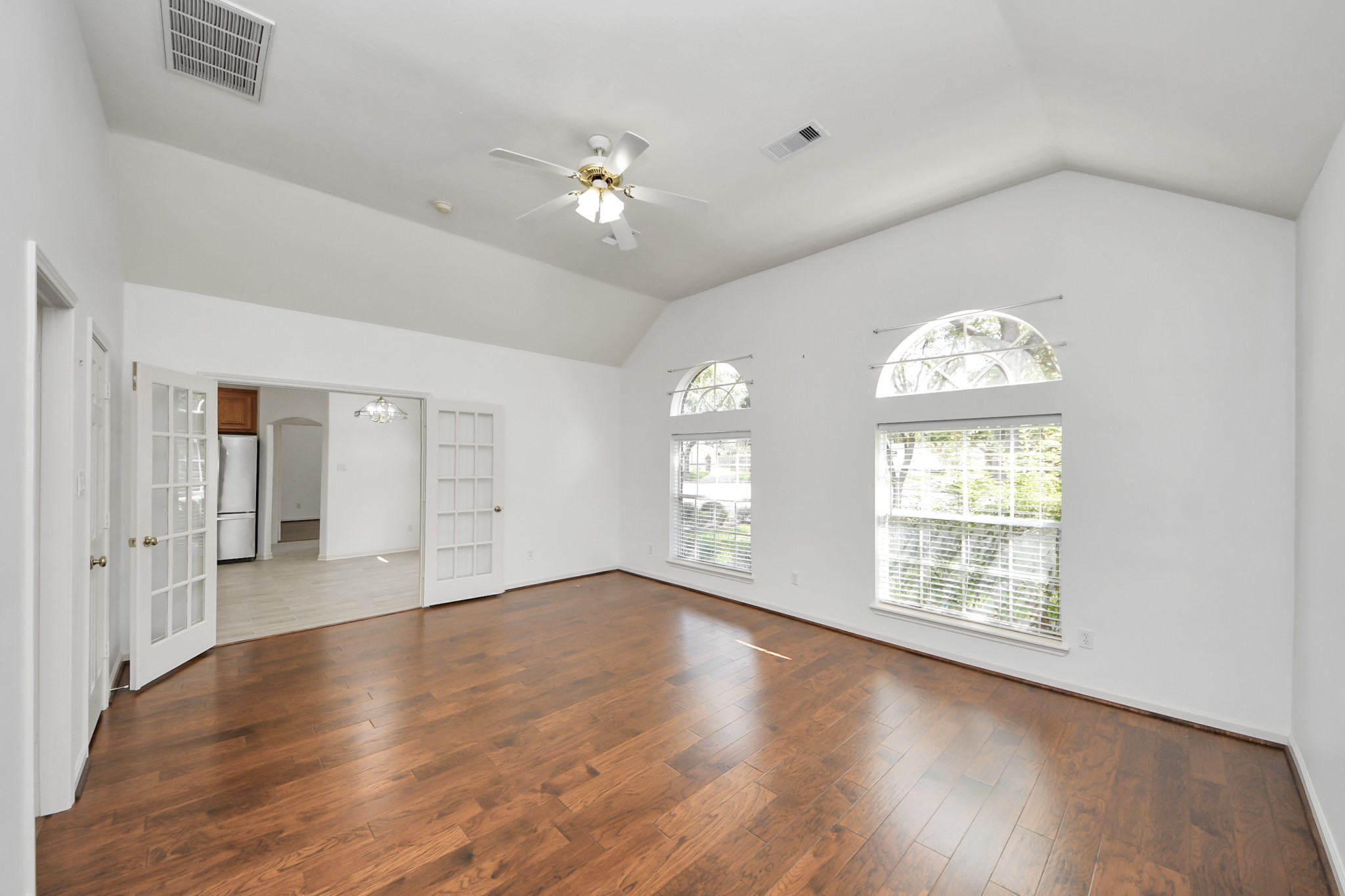 Spacious living area featuring elegant hardwood floors, abundant natural light from large arched windows, and a ceiling fan for comfort, seamlessly connected to the kitchen through stylish French doors.