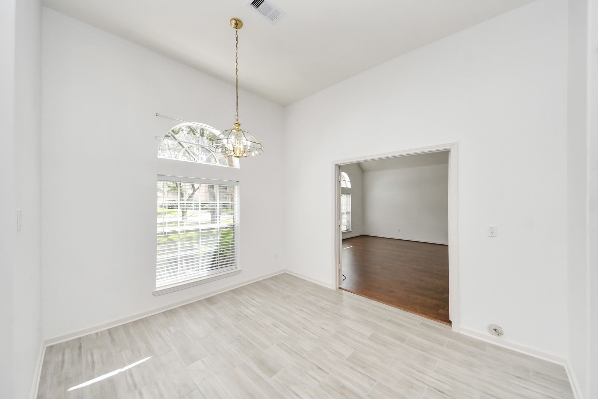 Bright and inviting living space featuring elegant tile flooring, a large arched window for natural light, and a stylish chandelier, seamlessly connecting to a cozy adjoining room with warm wood floors.