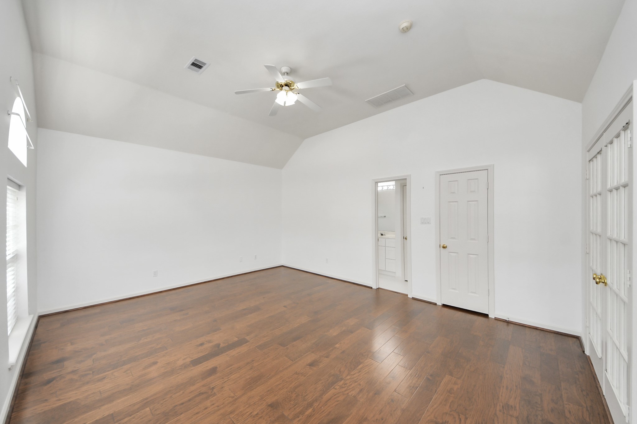 Spacious room with vaulted ceilings, elegant hardwood floors, and abundant natural light, featuring double French doors and convenient bathroom access.