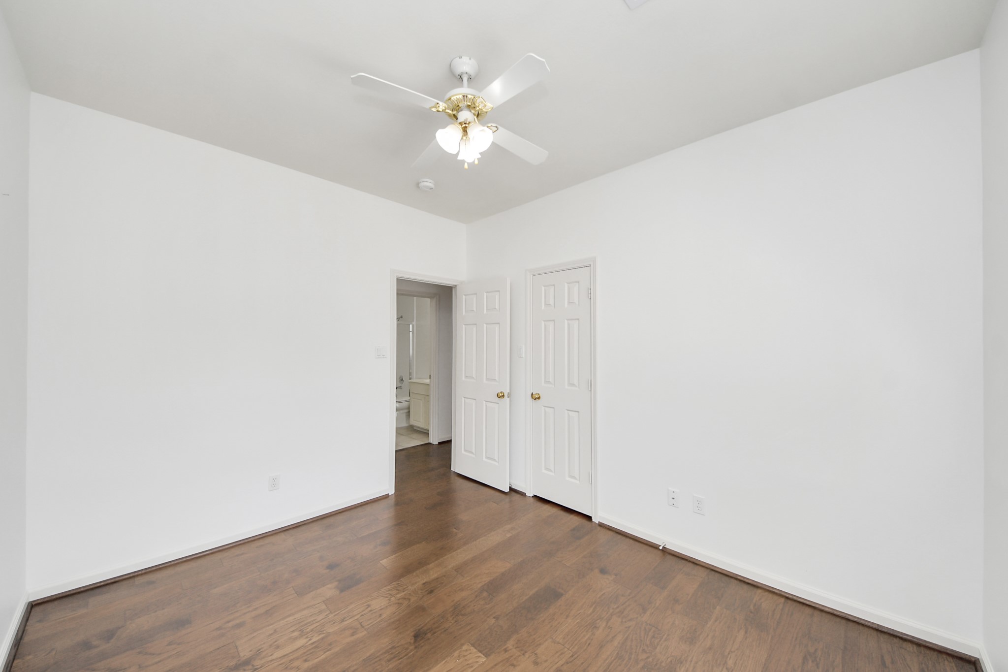 Bright room with elegant hardwood floors, a classic ceiling fan, and a seamless flow into an adjoining bathroom through double doors.