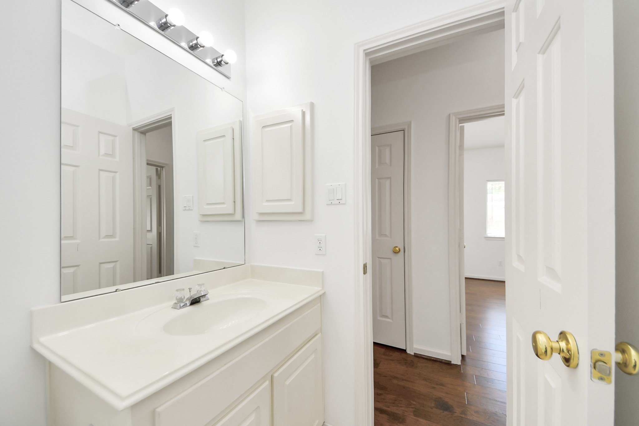 Bright bathroom features a sleek vanity with ample storage, modern lighting, and a large mirror, leading into a hallway with wood flooring.