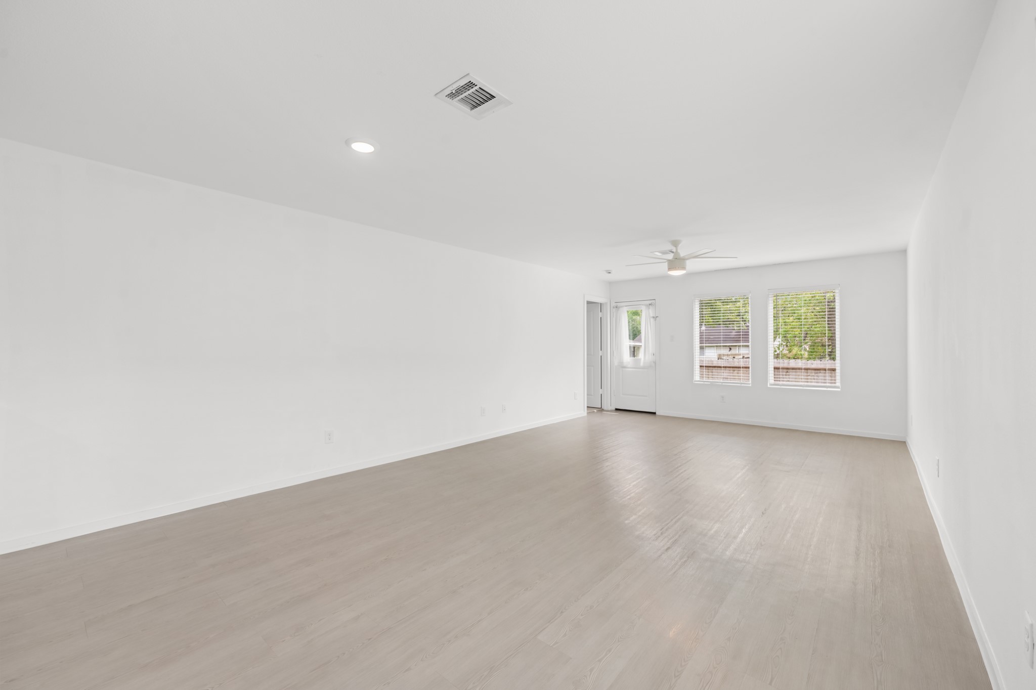 Spacious living area features light-toned flooring, neutral finishes, and a ceiling fan, with multiple windows bringing natural light into the space.