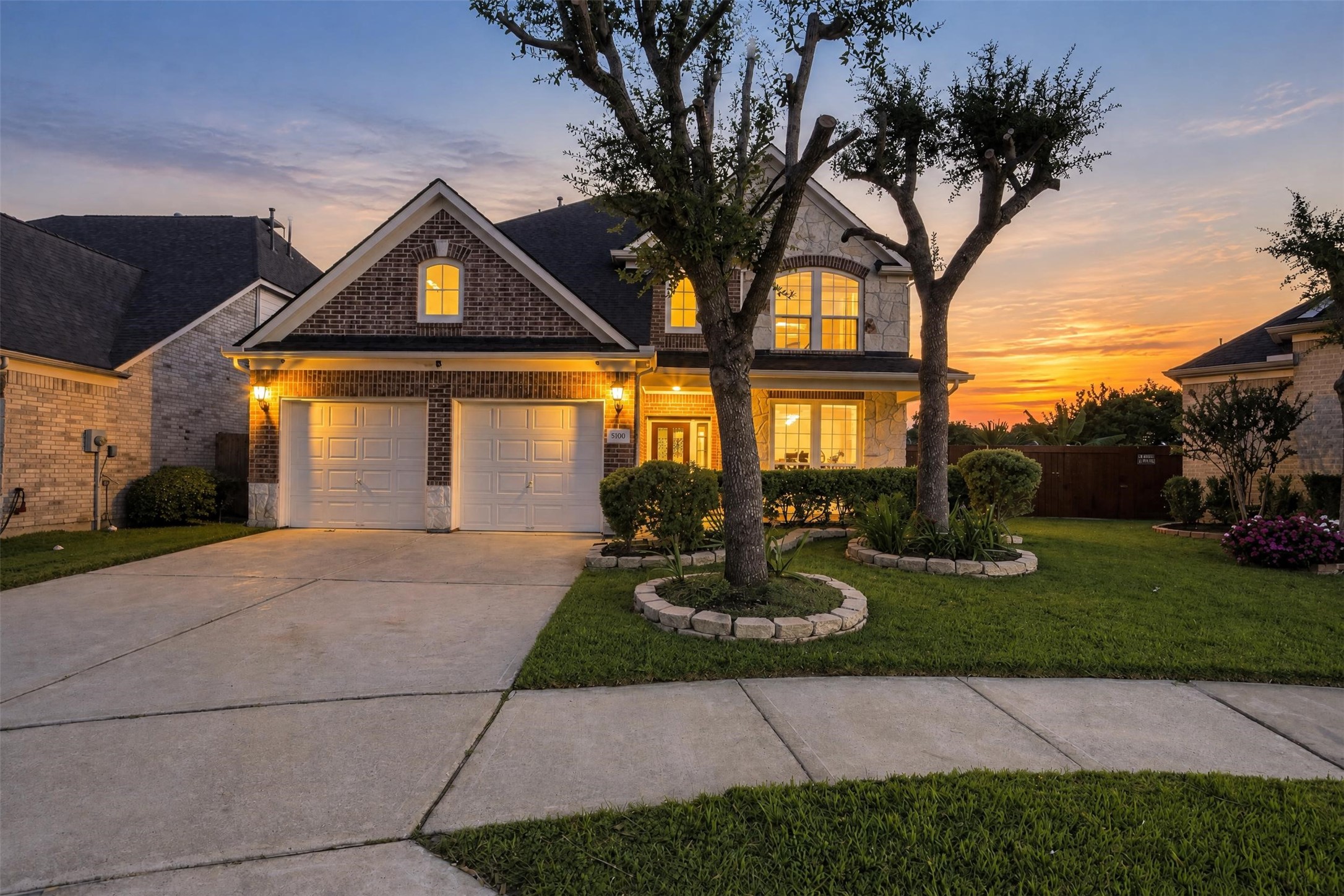 Welcome home at golden hour. Warm lights glow across the brick-and-stone front, mature trees frame the lawn, and the quiet cul-de-sac setting in Riverstone makes every evening arrival feel peaceful.