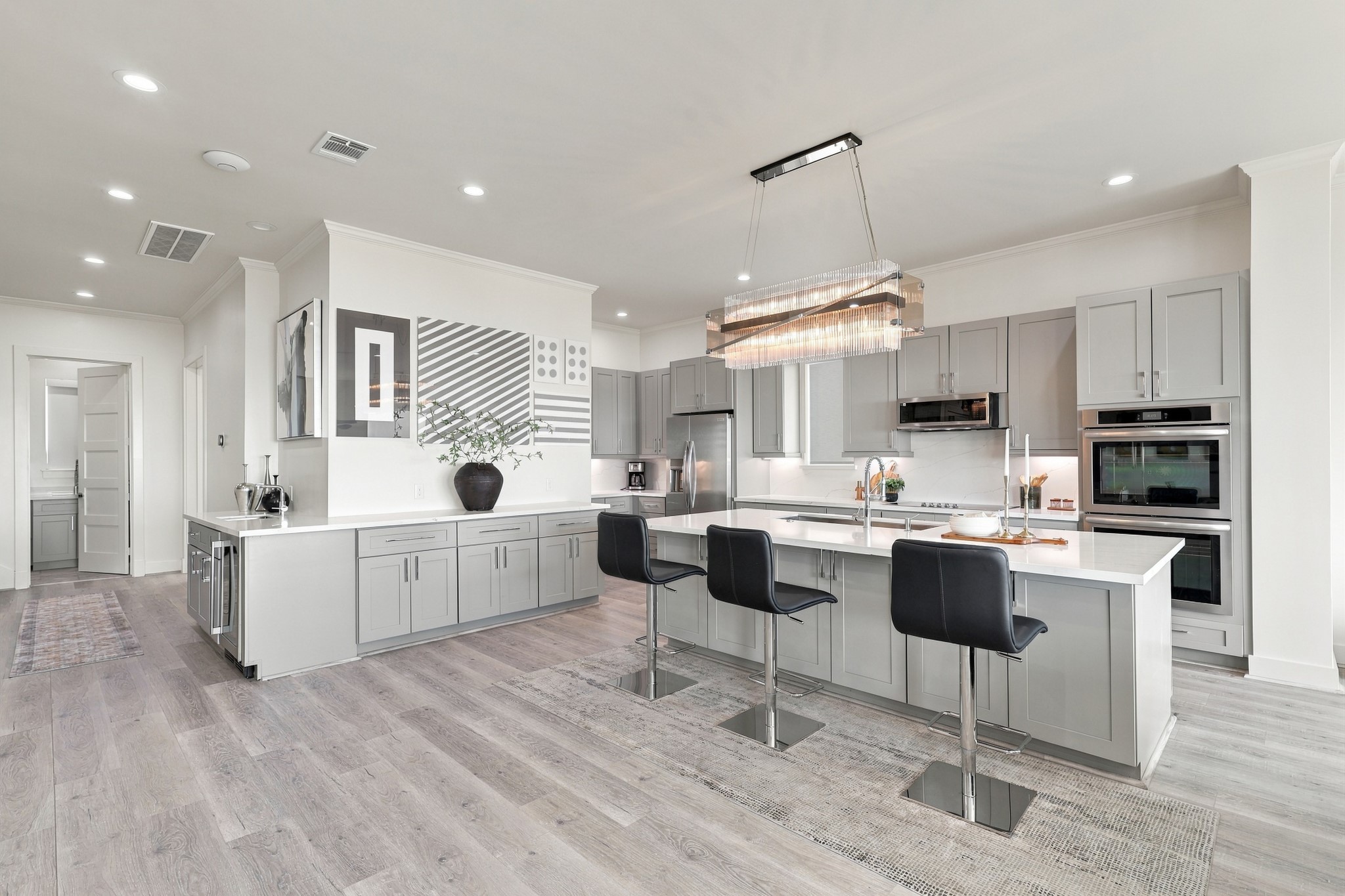 Sophisticated kitchen featuring gray cabinetry, expansive counters, stainless steel appliances, and double wall ovens. A statement chandelier highlights the oversized island with breakfast-bar seating.