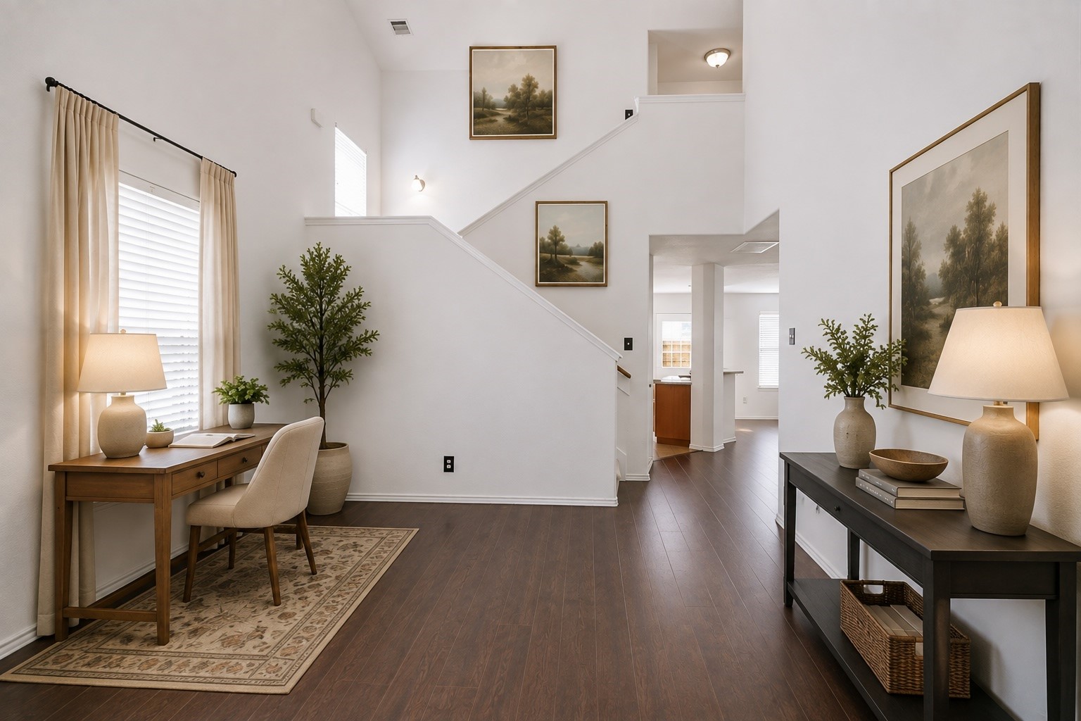 Soaring double-height ceilings elevate this open foyer, featuring rich wood-look flooring and a functional study nook tucked under the staircase. The bright space offers a seamless transition toward the main living areas and kitchen beyond. This photo has been virtually staged.