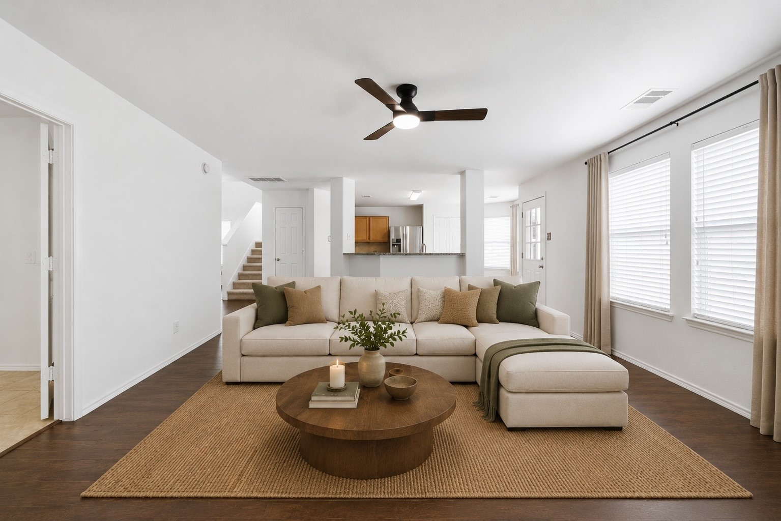 Abundant natural light pours into the open living area, showcasing dark wood-look floors, a ceiling fan, and crisp neutral walls. Seamless sightlines lead straight into the kitchen and adjacent staircase, creating an inviting layout. This photo has been virtually staged.