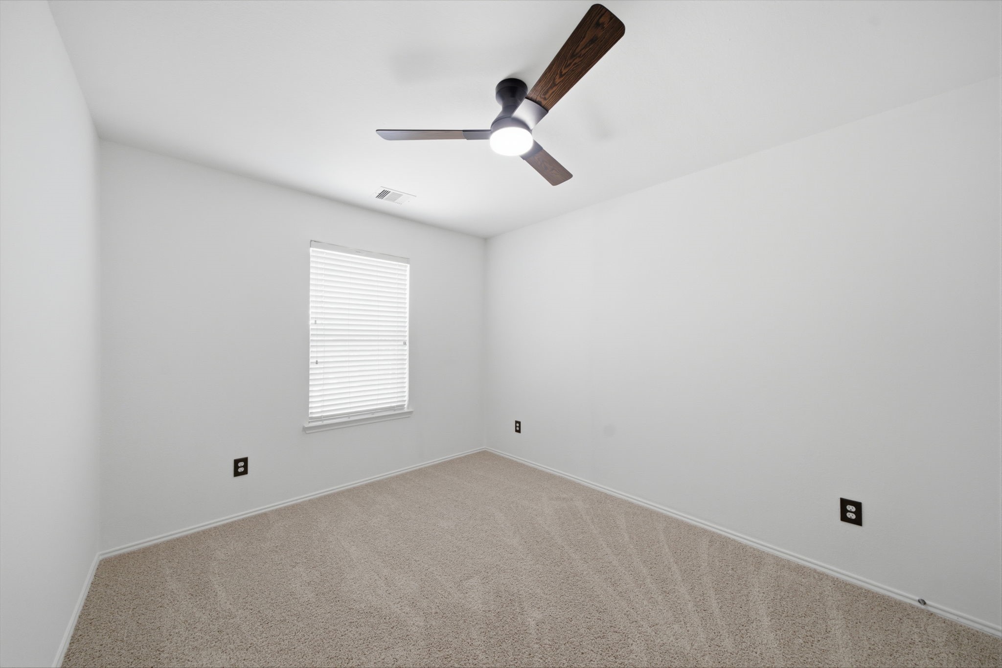 Plush carpet flooring and a dark-blade ceiling fan complement the fresh neutral paint in this secondary bedroom. A single window brings in natural light, framing a clean, adaptable space for rest or study.