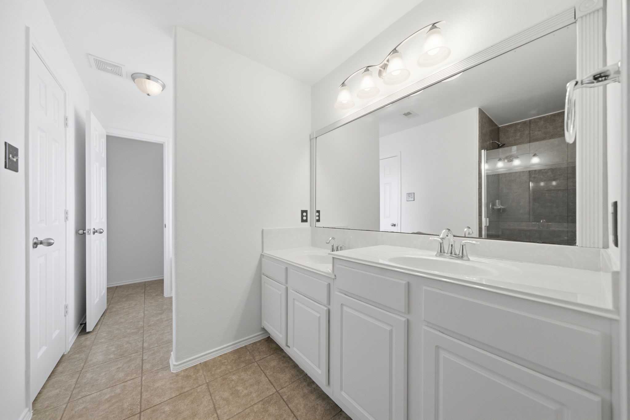 A extended white vanity with dual sinks and chrome fixtures highlights this primary bath, offering generous cabinet storage and a wide mirror. A separate glass-enclosed shower with tile surrounds and a nearby water closet ensure privacy and routine efficiency.
