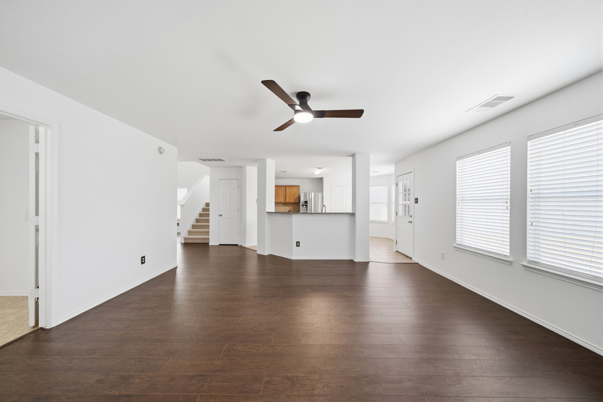 Dark wood-look flooring and crisp neutral walls anchor this spacious first-floor living area, featuring a modern ceiling fan and direct sightlines to the kitchen's breakfast bar. Large windows fill the open layout with light, while convenient access to the staircase and secondary rooms creates an effortless flow.