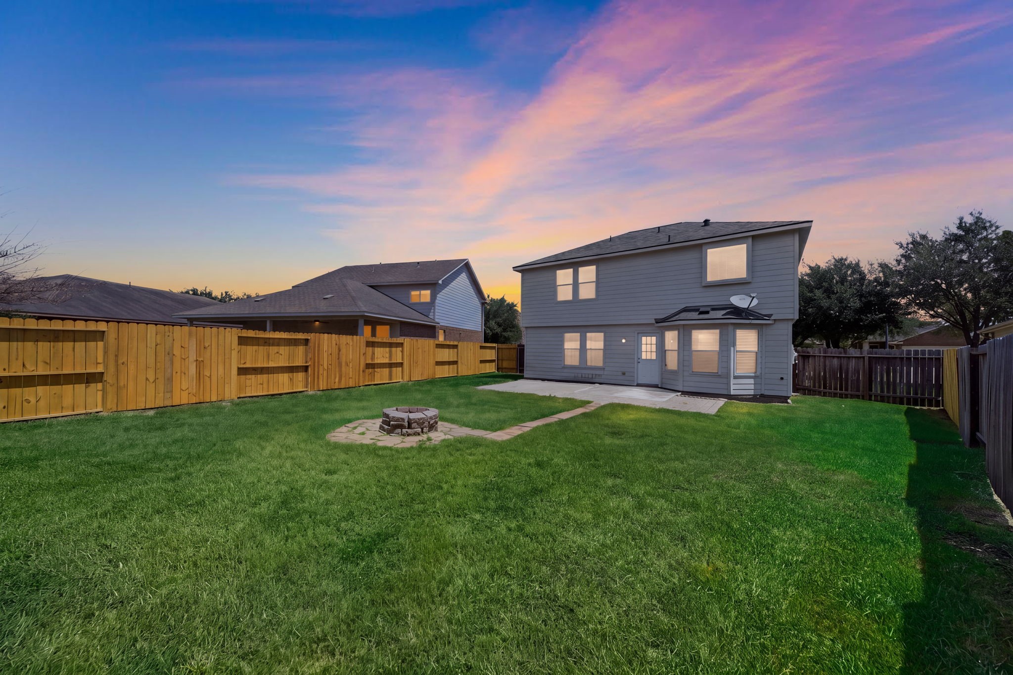 A stone fire pit with a flagstone path serves as the focal point in this fully fenced, expansive green backyard featuring a concrete patio.