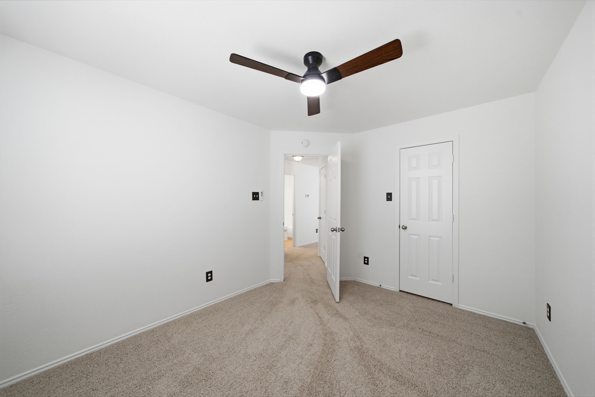 Soft neutral carpet and a modern dark-blade ceiling fan define this secondary bedroom featuring fresh white paint. A full-size closet door sits beside the entry door, framing an efficient layout for guests or a home office.