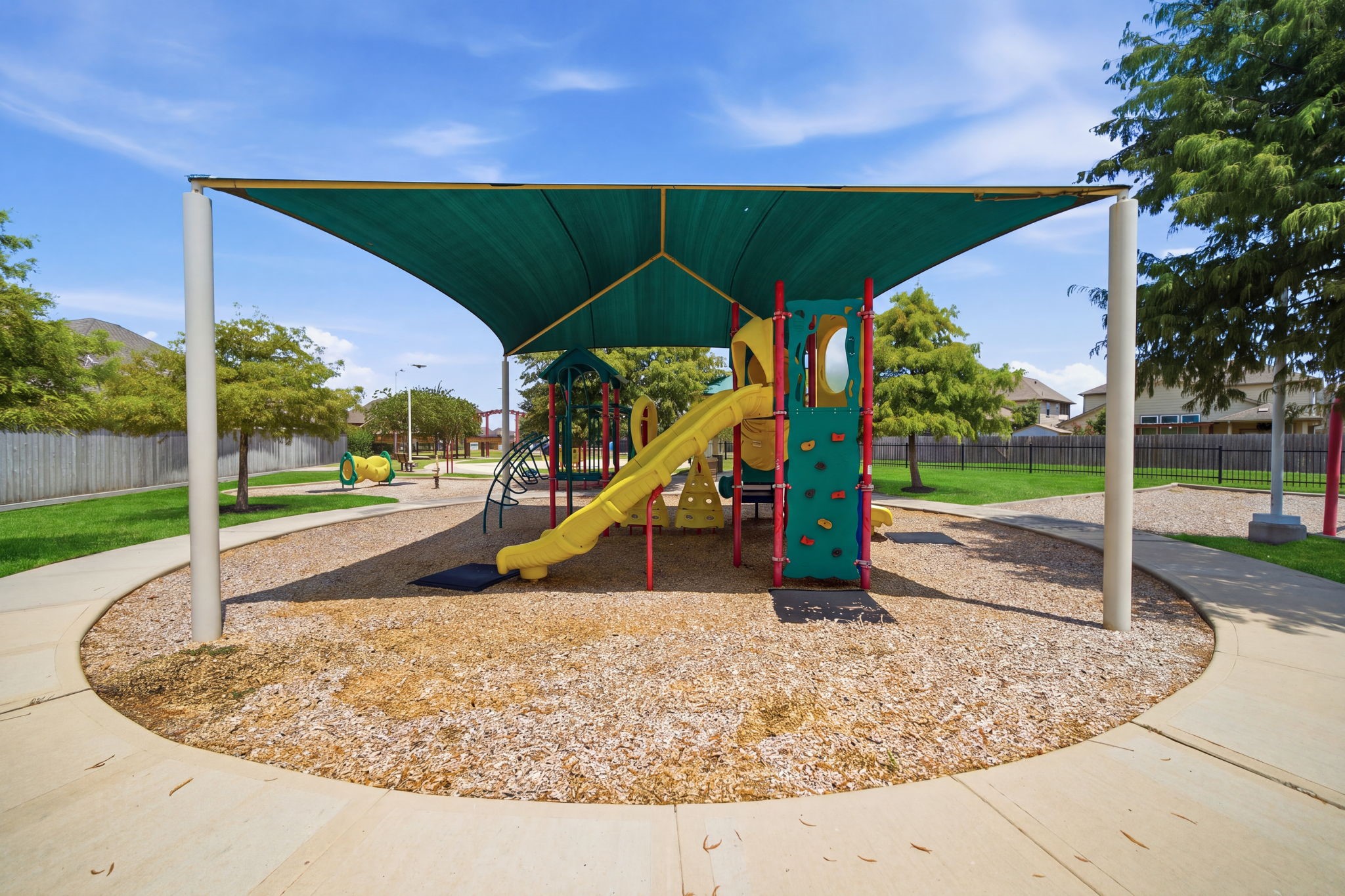 A shade canopy covers this neighborhood playground featuring a slide, climbing wall, and surrounding walking path.