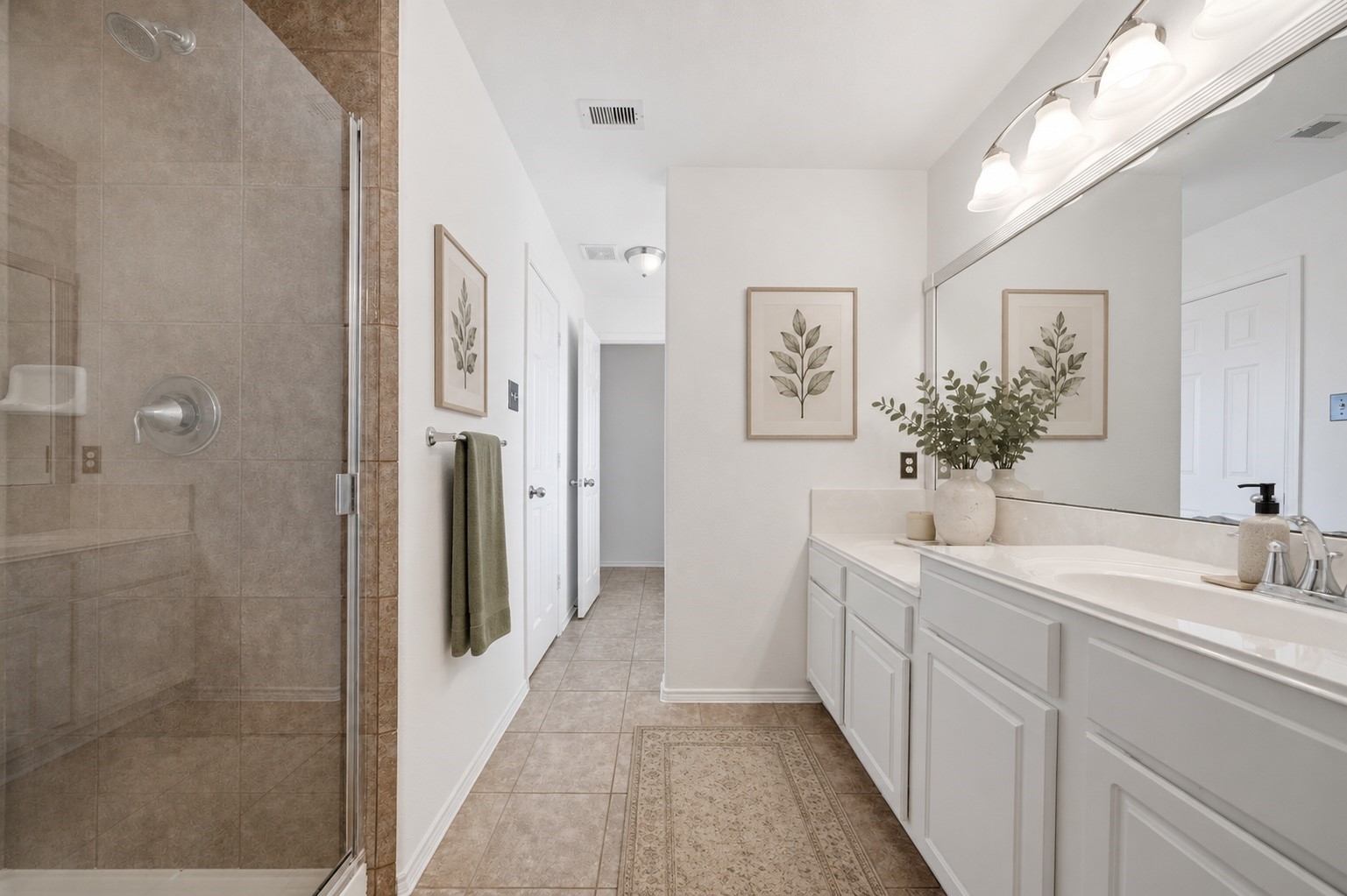 Crisp white vanities with dual sinks extend along tile flooring, offering ample counter space and storage beneath a large framed mirror. A glass-enclosed shower featuring neutral tile surrounds completes this spacious ensuite bath.