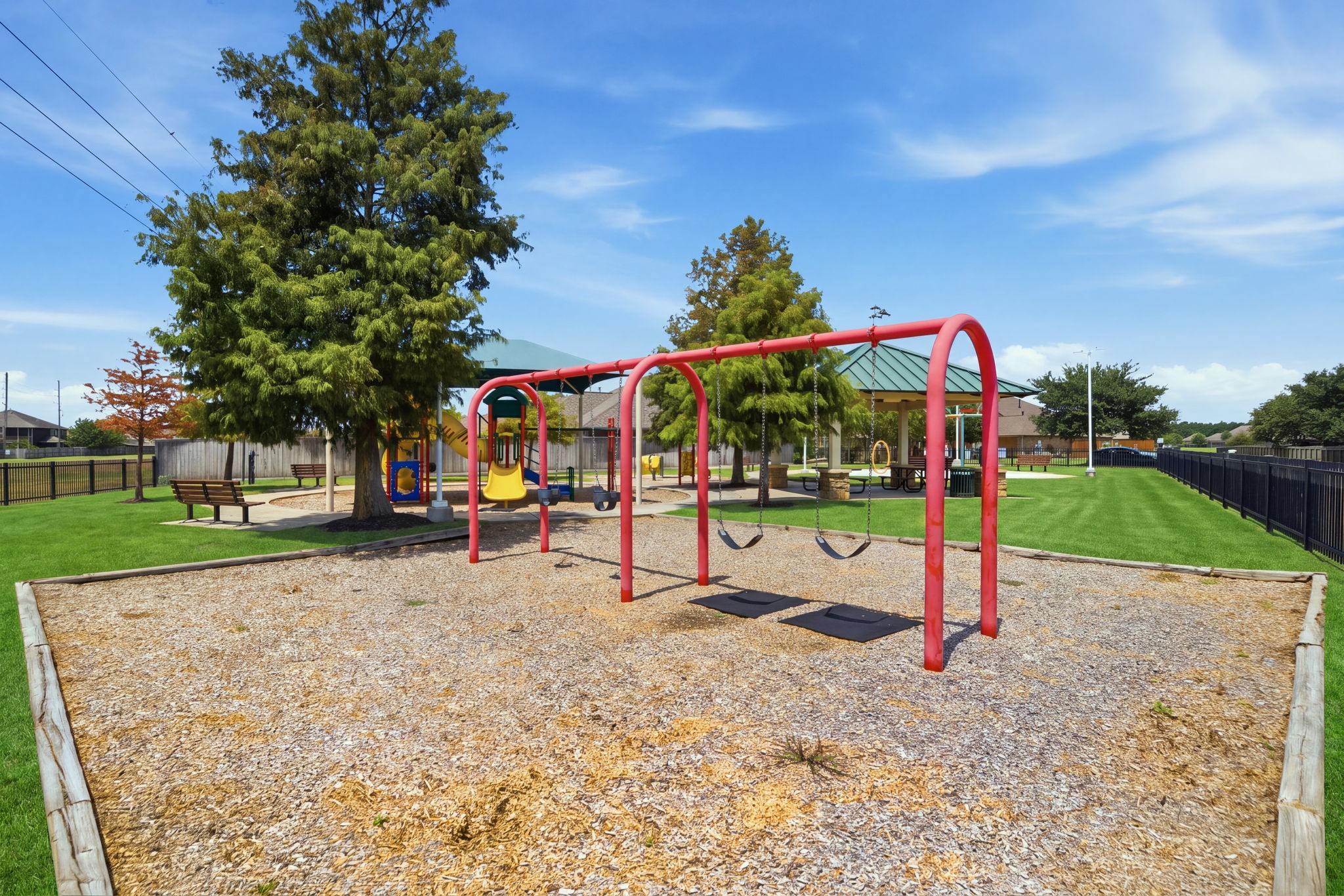 A red swing set anchors this neighborhood playground, featuring a wood-chip surface, swings, and a background pavilion with green space.