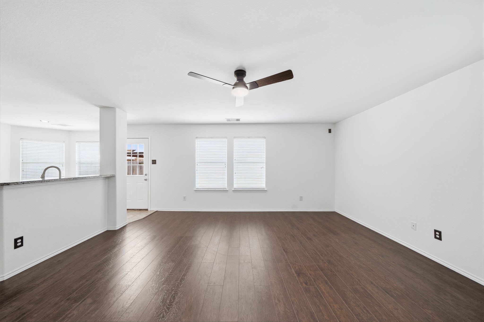 Gleaming dark wood-look floors span this first-floor living space, complete with a modern ceiling fan and large windows with blinds. An open wall cut-out provides a direct view into the kitchen, ensuring effortless interaction across main rooms.