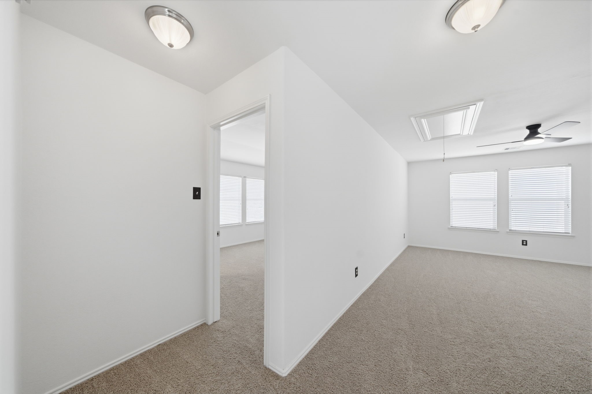 Neutral carpet flooring and crisp white walls anchor this second-floor hallway connecting the upstairs bedrooms and gameroom. A ceiling fan and flush-mount fixtures illuminate the open layout, while a ceiling hatch provides convenient access to attic storage.