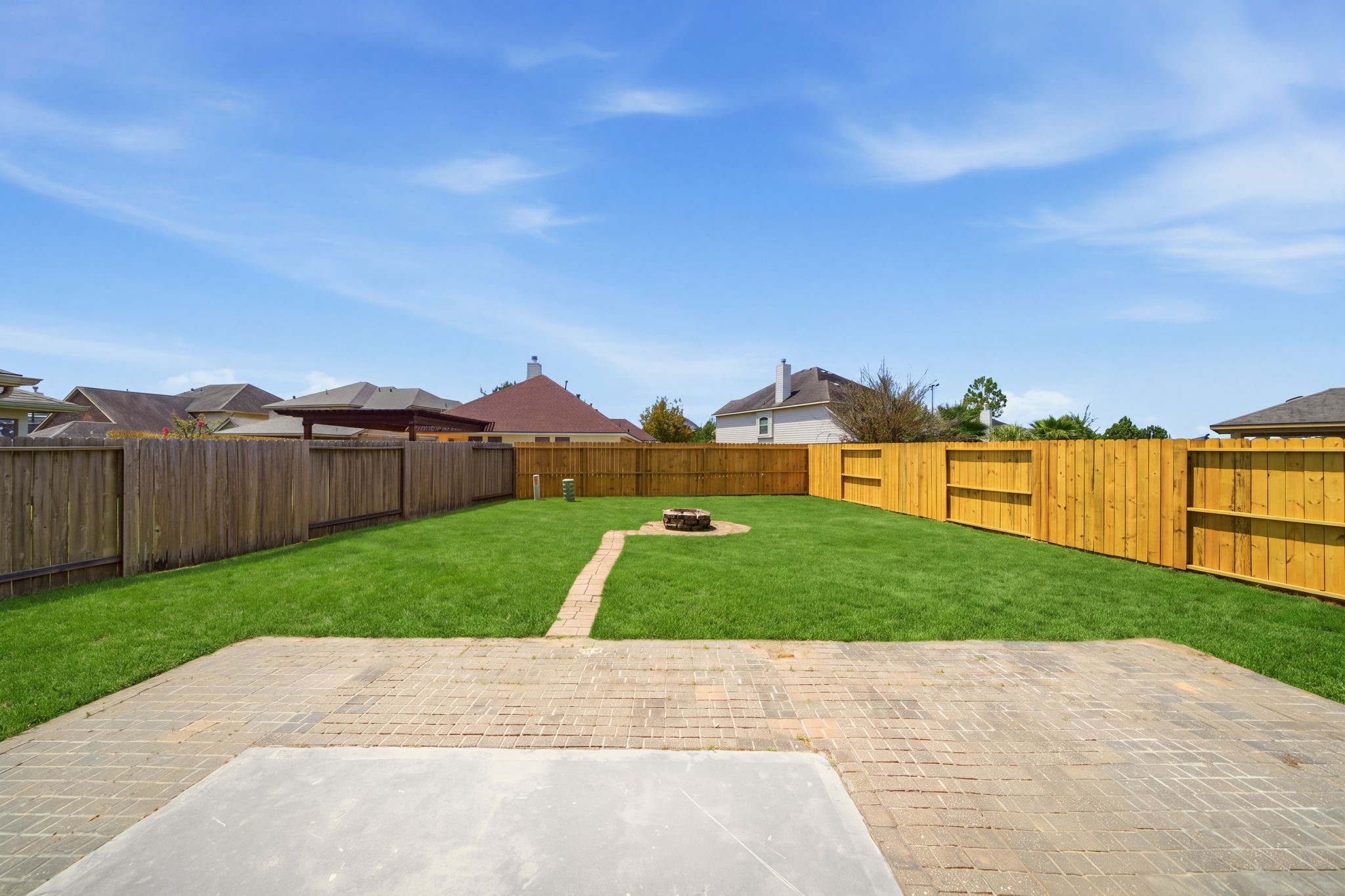 A brick paver patio extends into a vast green lawn within this private fenced yard, featuring a paved walkway that leads to a central fire pit. Plenty of open space provides endless possibilities for a custom pool or a lush garden setup.