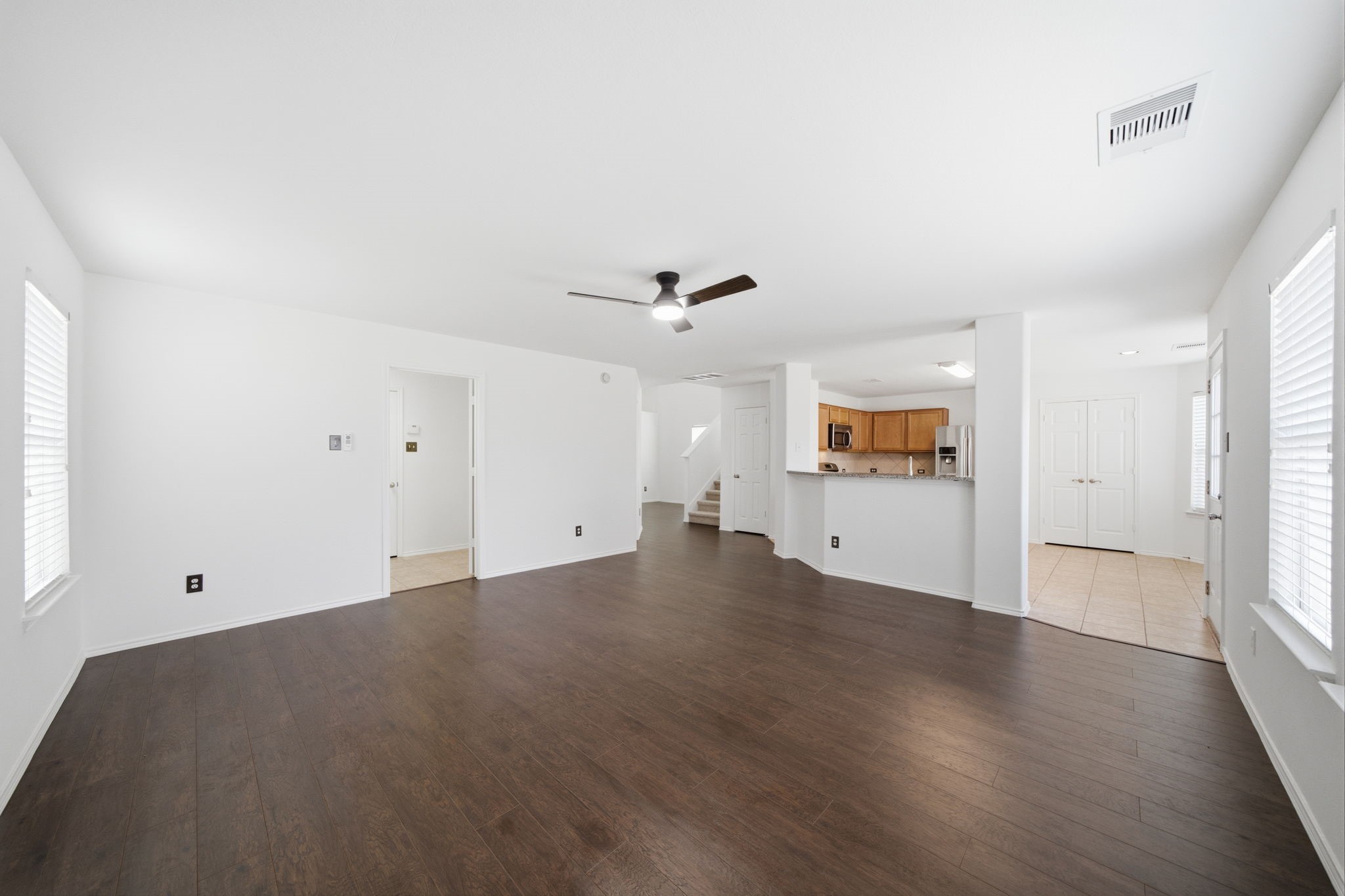 Rich dark hardwood floors and crisp white walls frame this spacious first-floor living area featuring a central ceiling fan. An open layout connects the space directly to the kitchen's breakfast bar, while wide windows provide ample light.