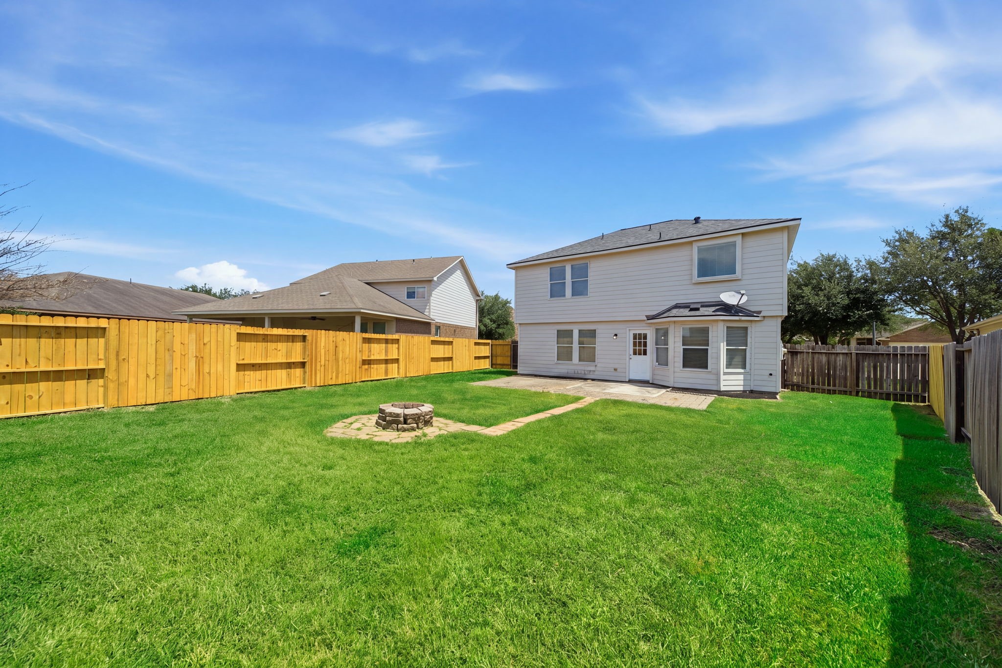 A expansive green lawn surrounds a stone fire pit, offering a private fenced outdoor space with a concrete patio.