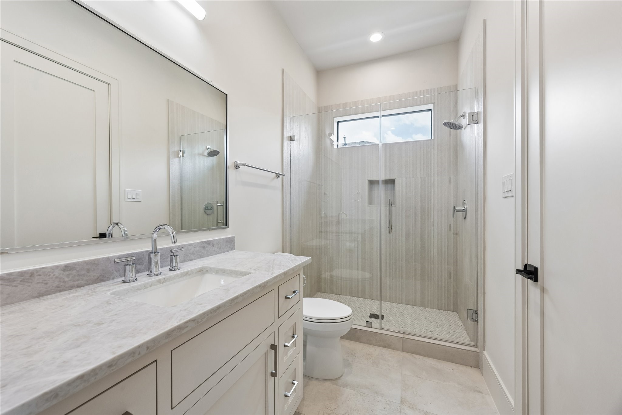 Soft neutral finishes, a quartz vanity, and a frameless glass shower create a timeless ensuite bath. Full height tile, a built in shower niche, and transom window add both style and natural light.