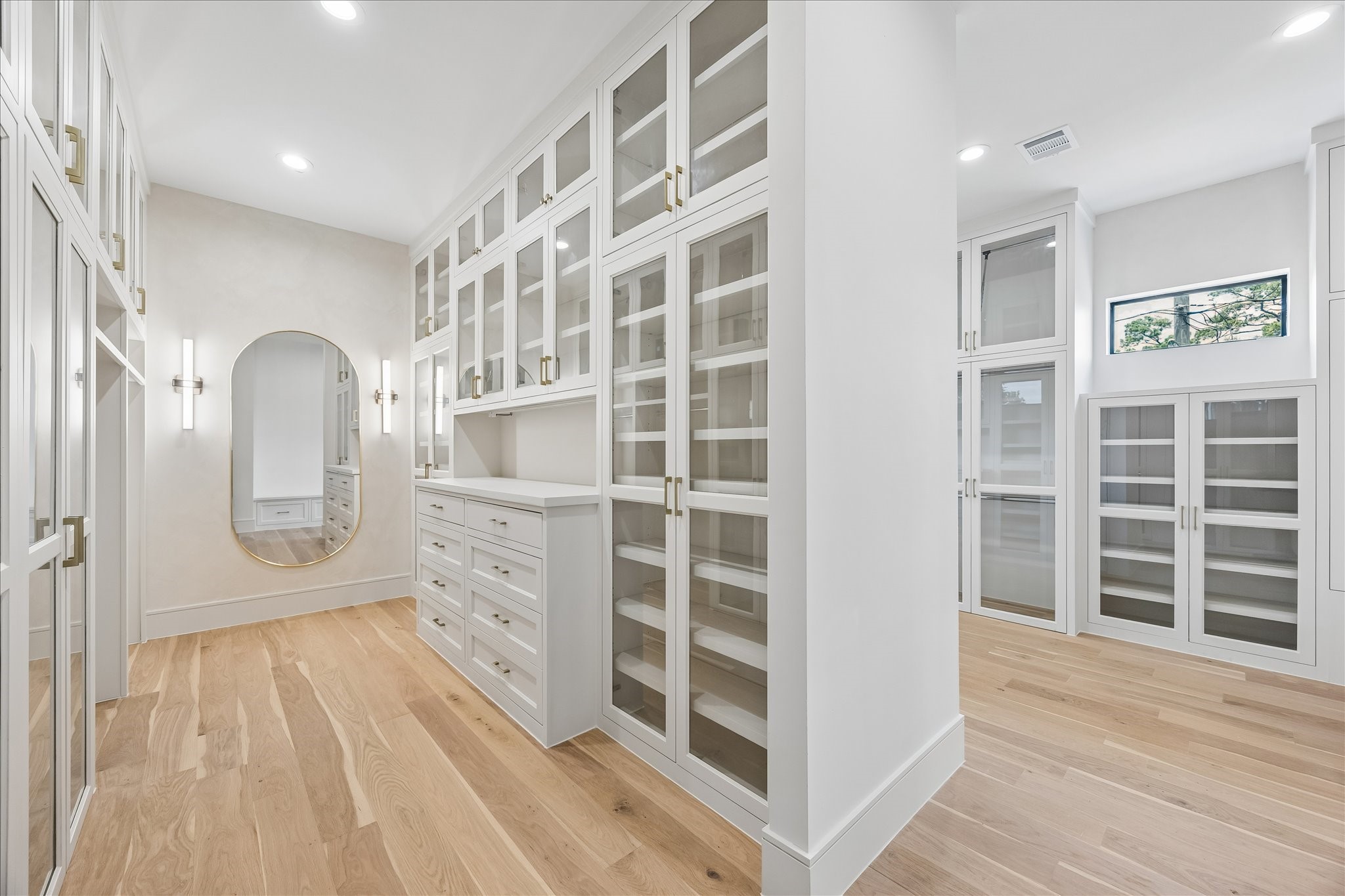 Boutique style dressing room featuring floor to ceiling custom cabinetry, glass display doors, integrated drawer storage, and a center dressing station. The back wall is lime washed to match the homes soft white palette and finished with designer sconces and mirrors, with all cabinetry pre-wired for LED lighting to be added as an upgrade. Thoughtfully designed with abundant storage, natural light, and direct access to the spa inspired primary bath.
