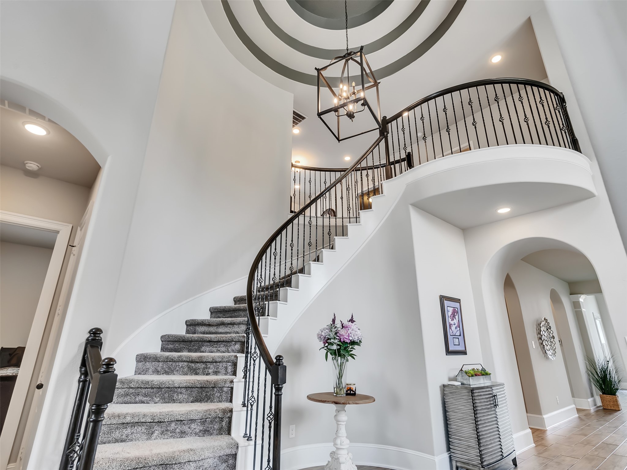 A curved staircase with iron railings, a high ceiling with a stylish light fixture, and neutral tones. A curved staircase with iron railings, a high ceiling with a stylish light fixture, and neutral tones.