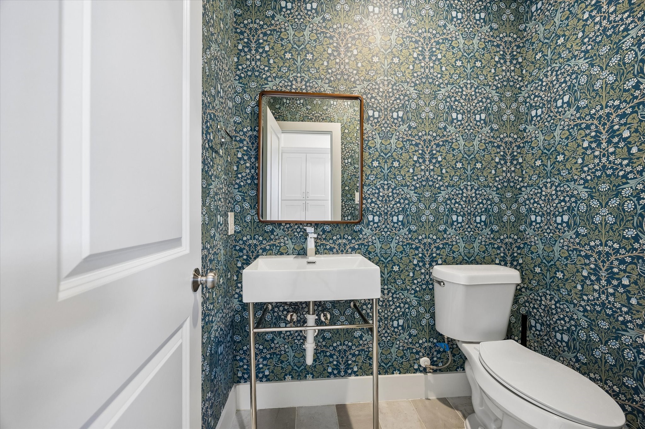 This chic powder room located in the mud room, features a sleek pedestal sink with a modern faucet and a framed mirror. The bold wallpaper creates a bright and inviting space.
