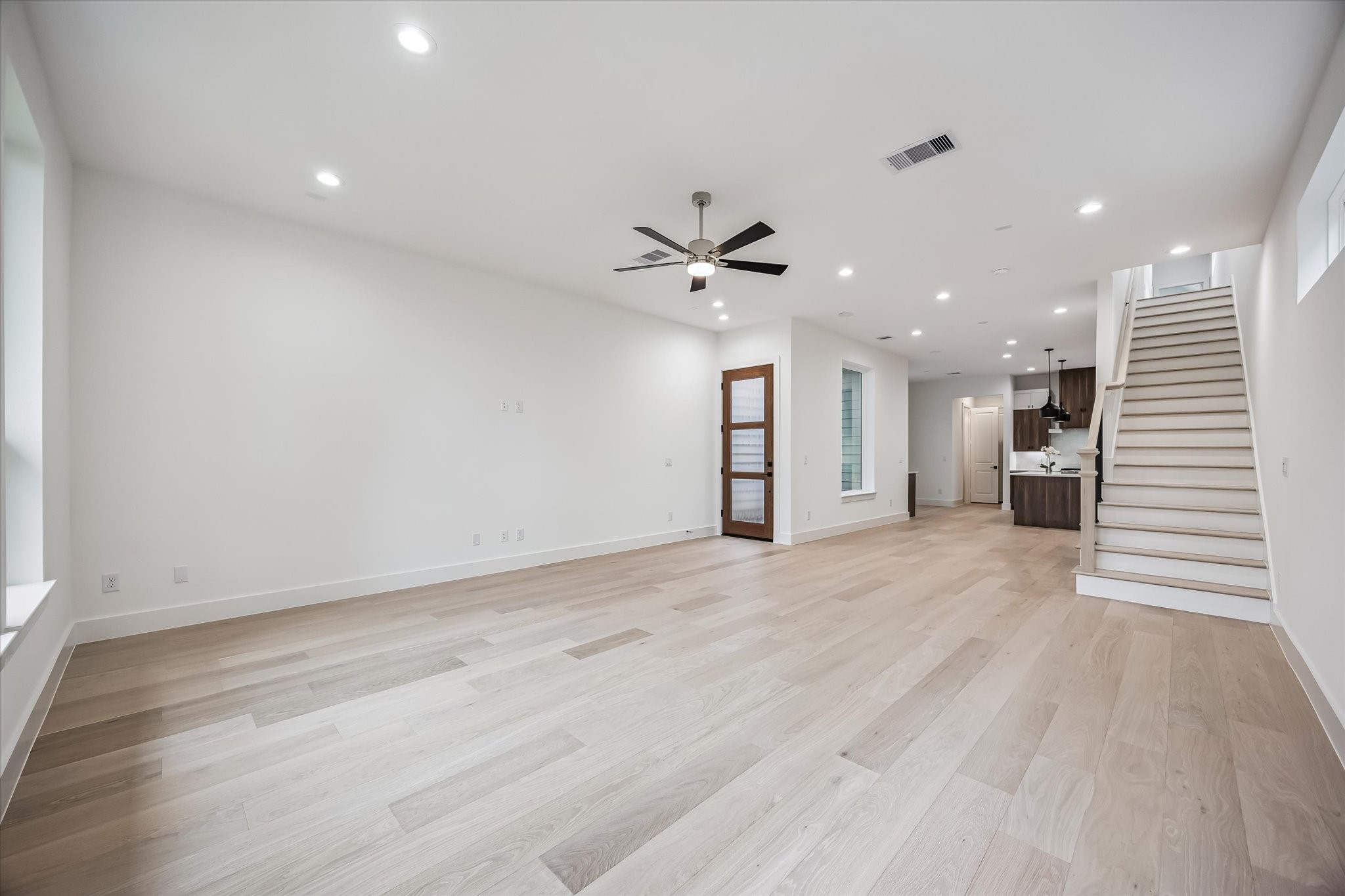 Spacious and bright living area featuring elegant wood flooring, recessed lighting, and a modern ceiling fan, seamlessly leading to a contemporary kitchen and staircase.
