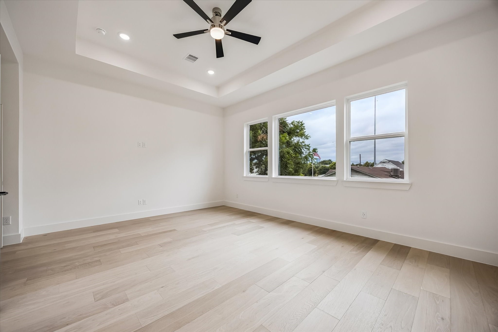 Bright and spacious room featuring elegant hardwood floors, large windows for natural light, and a modern ceiling fan set within a chic tray ceiling.