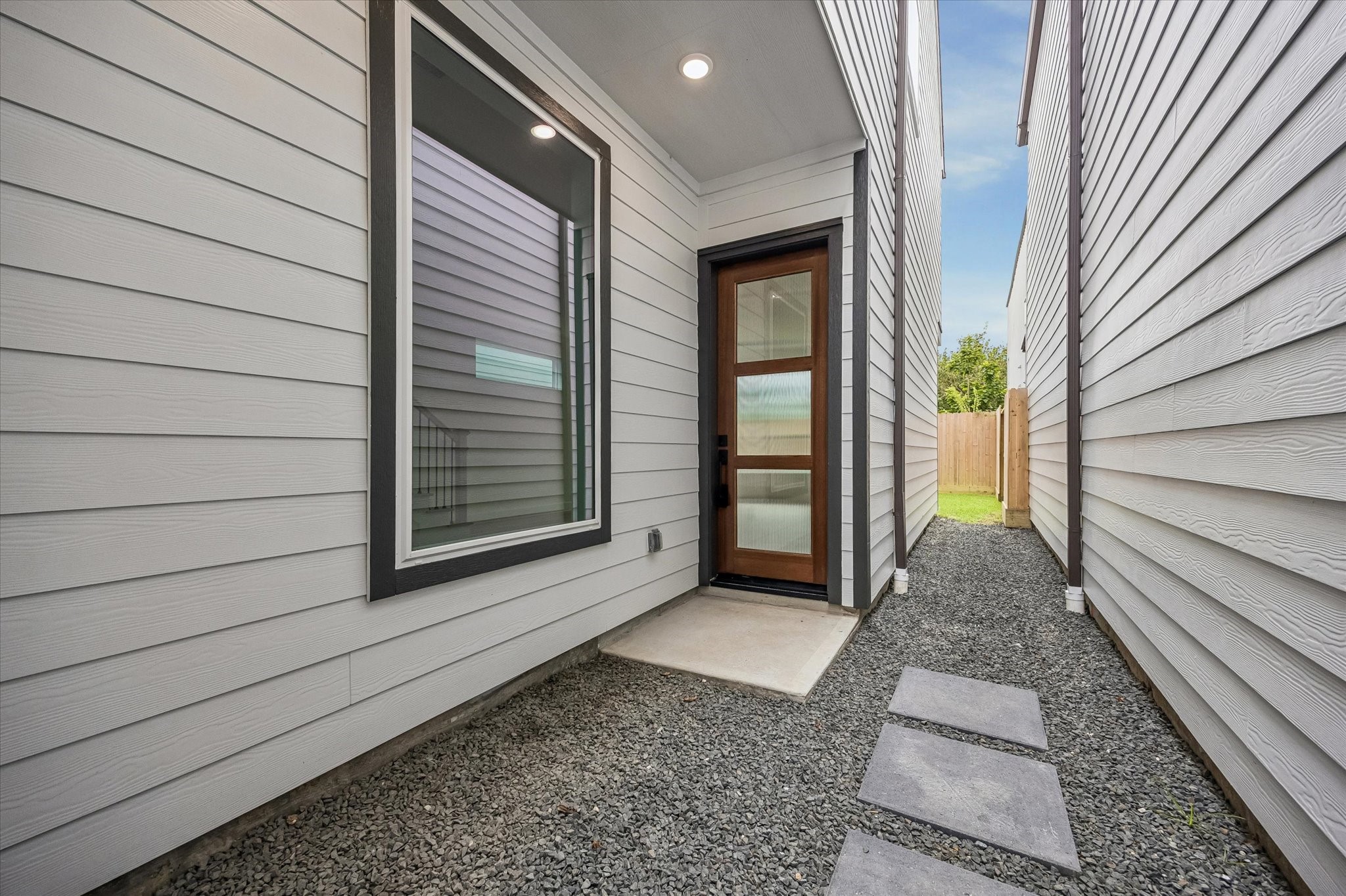 Modern side entrance with sleek wood and glass door, framed by clean white siding, leading to a private backyard space.