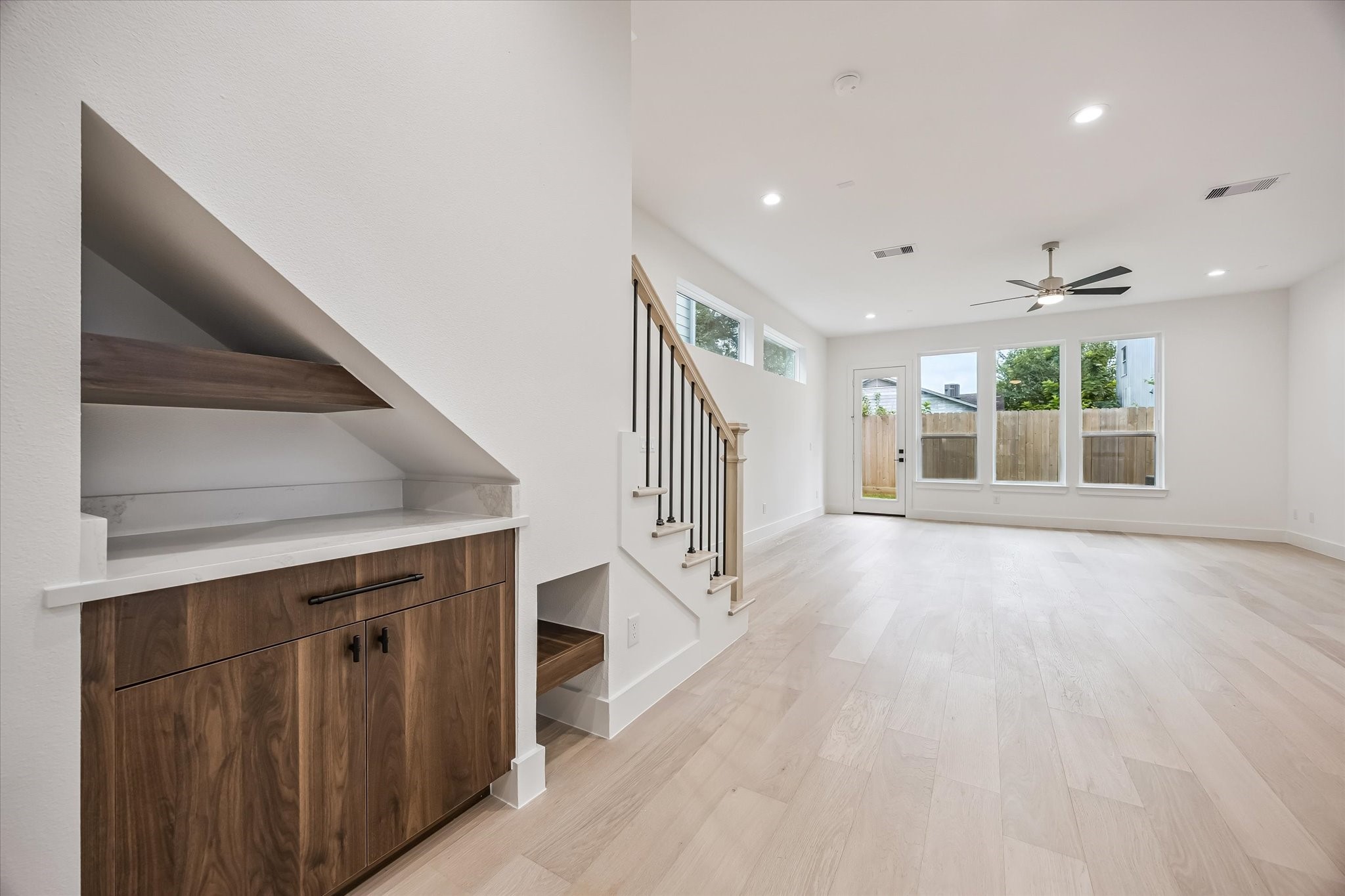 Bright and spacious living area featuring sleek hardwood floors, a modern ceiling fan, and large windows that flood the room with natural light, complemented by a stylish built-in cabinet under the staircase.