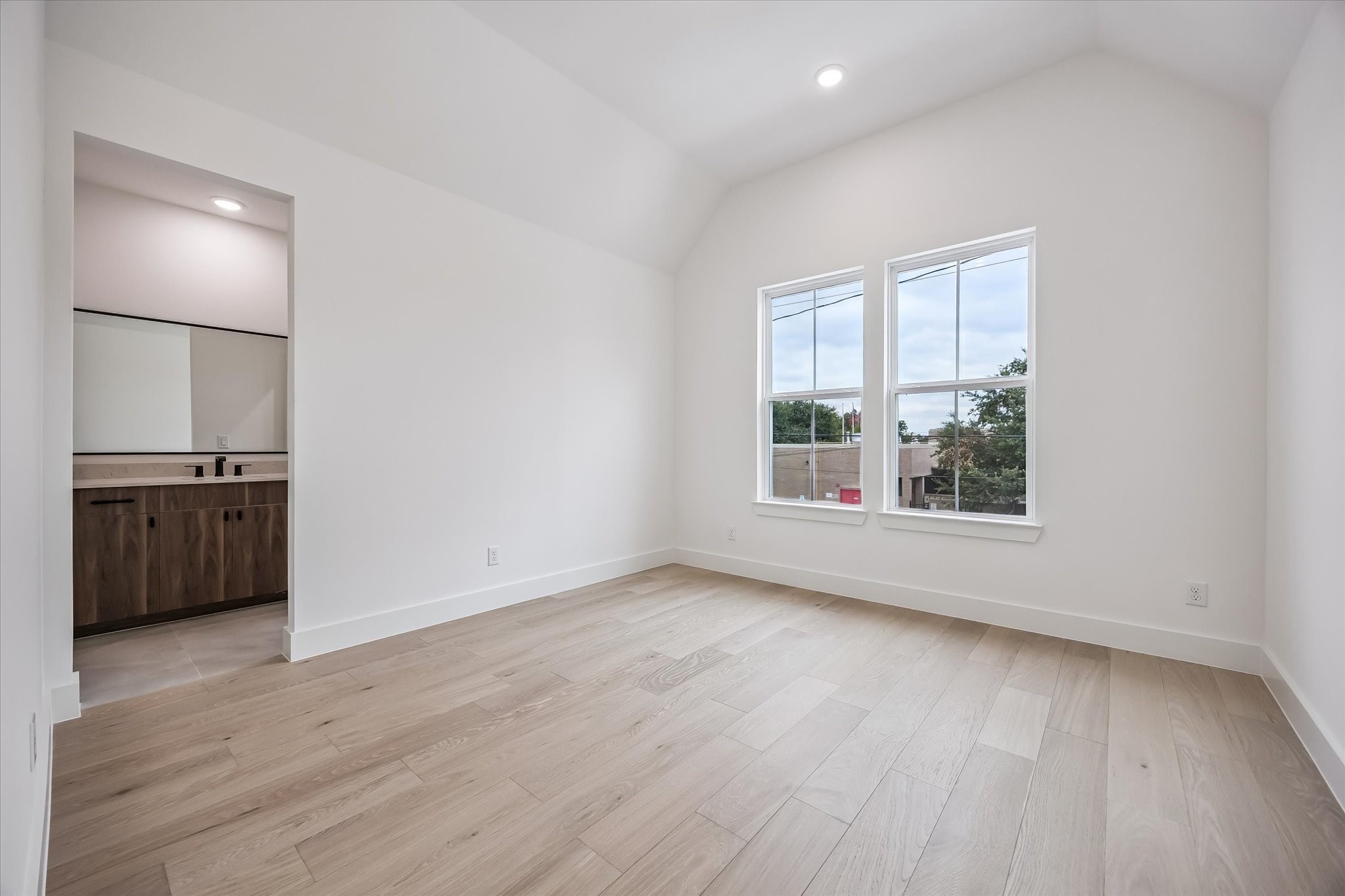 Bright and airy bedroom with vaulted ceilings, wood flooring, and large windows, leading into a modern en-suite bathroom with sleek cabinetry.