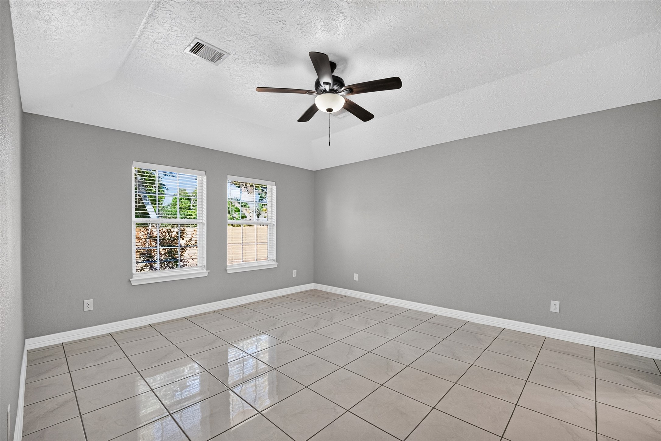 Spacious primary bedroom with sleek tile flooring, neutral gray walls, and ample natural light from two large windows, complemented by a modern ceiling fan and en suite.