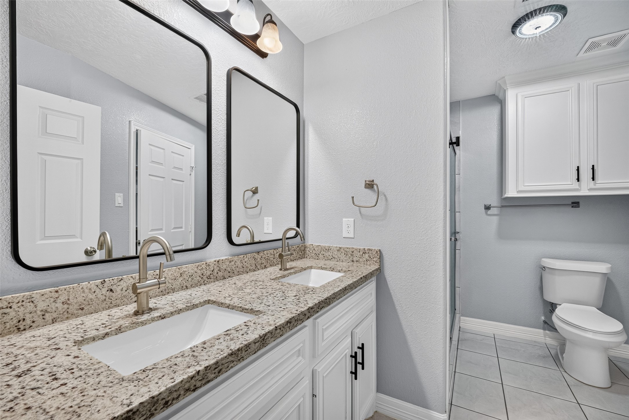 Elegant en suite featuring a dual-sink vanity with granite countertops, modern fixtures, and ample storage, complemented by soft gray walls and tile flooring.