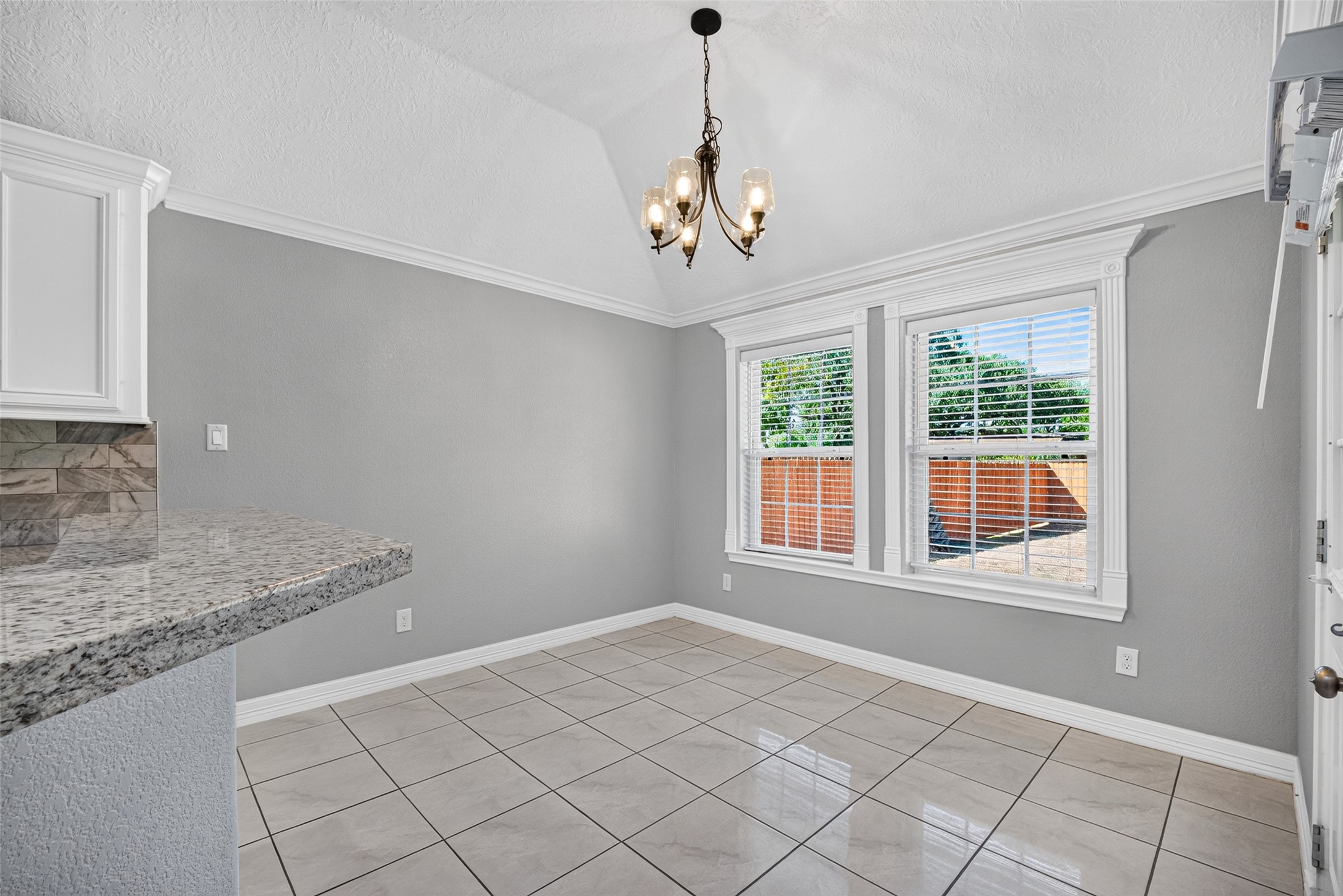 Bright dining area with vaulted ceiling, elegant chandelier, and large windows offering plenty of natural light. Stylish tile flooring and a sleek granite breakfast bar add a modern touch.
