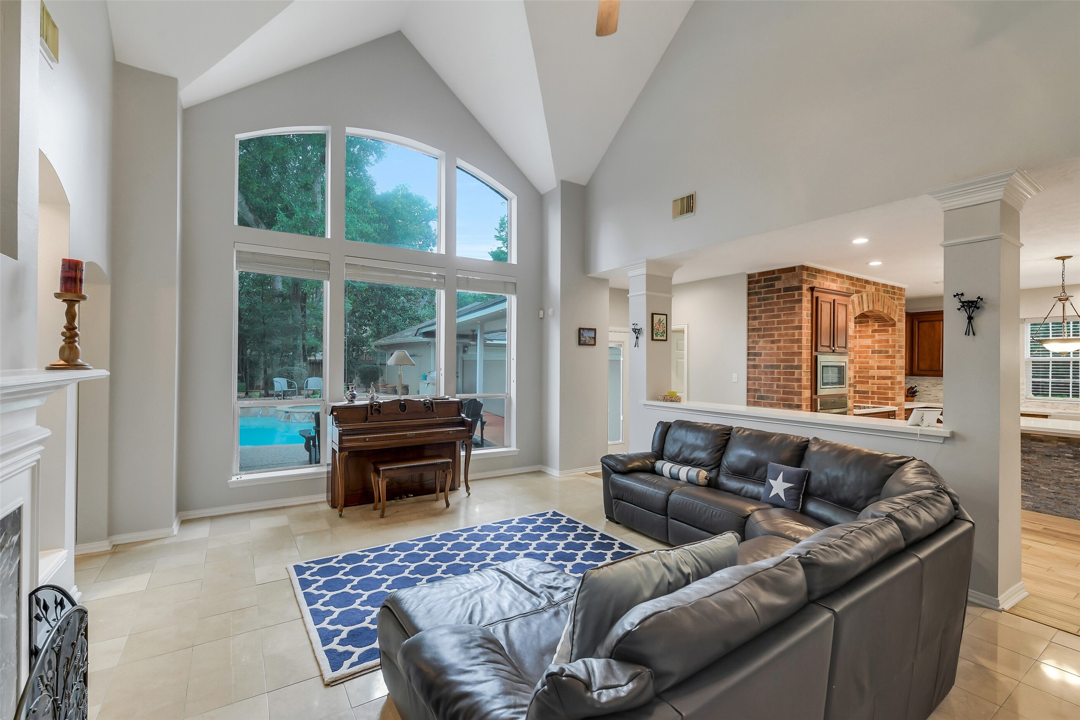 Spacious living area with soaring vaulted ceilings, large windows overlooking the pool, and a cozy fireplace, seamlessly connected to a modern kitchen with brick accents.