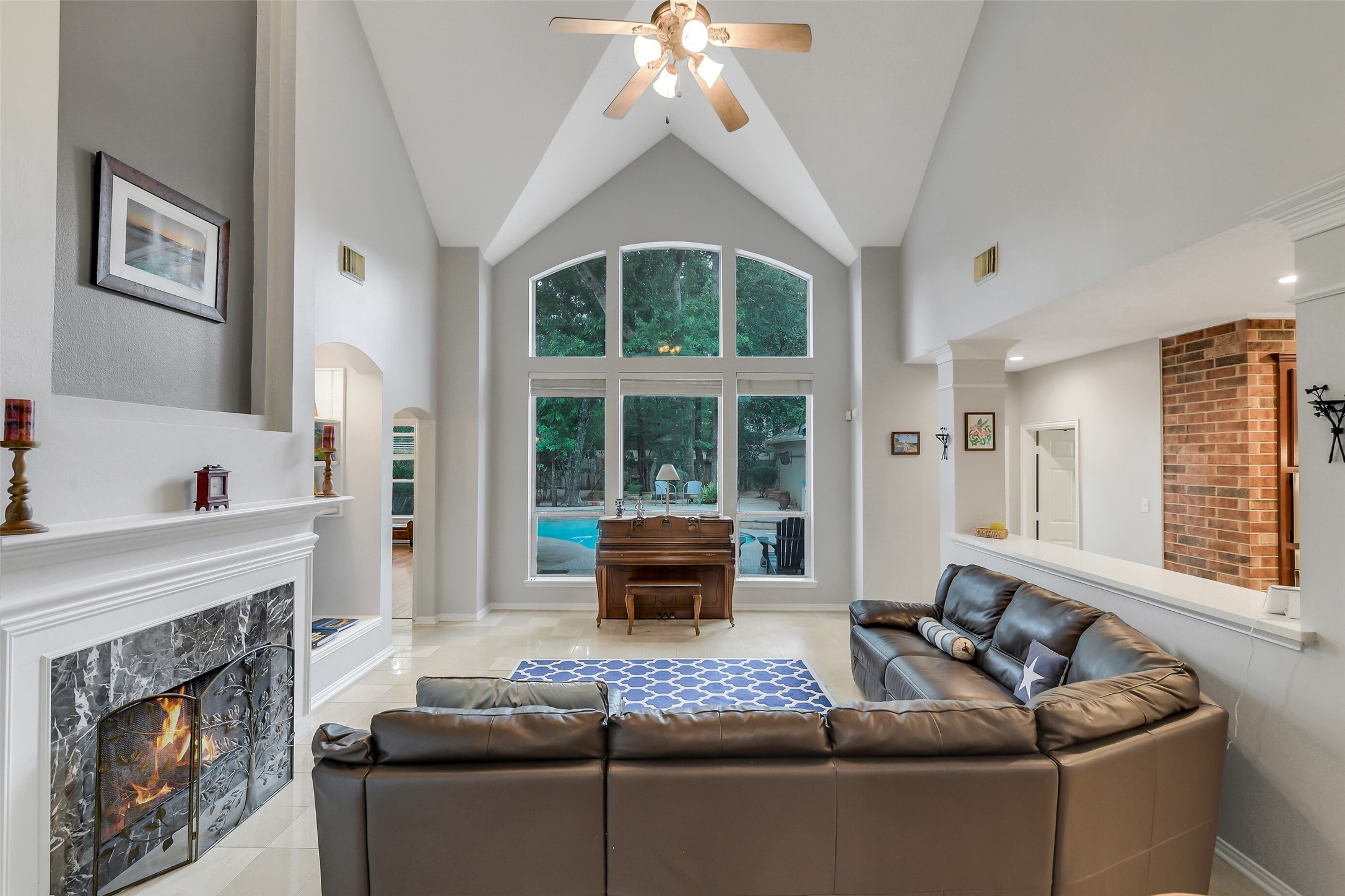 Spacious living room with vaulted ceilings, a cozy fireplace, and large arched windows offering serene views of the backyard and pool.