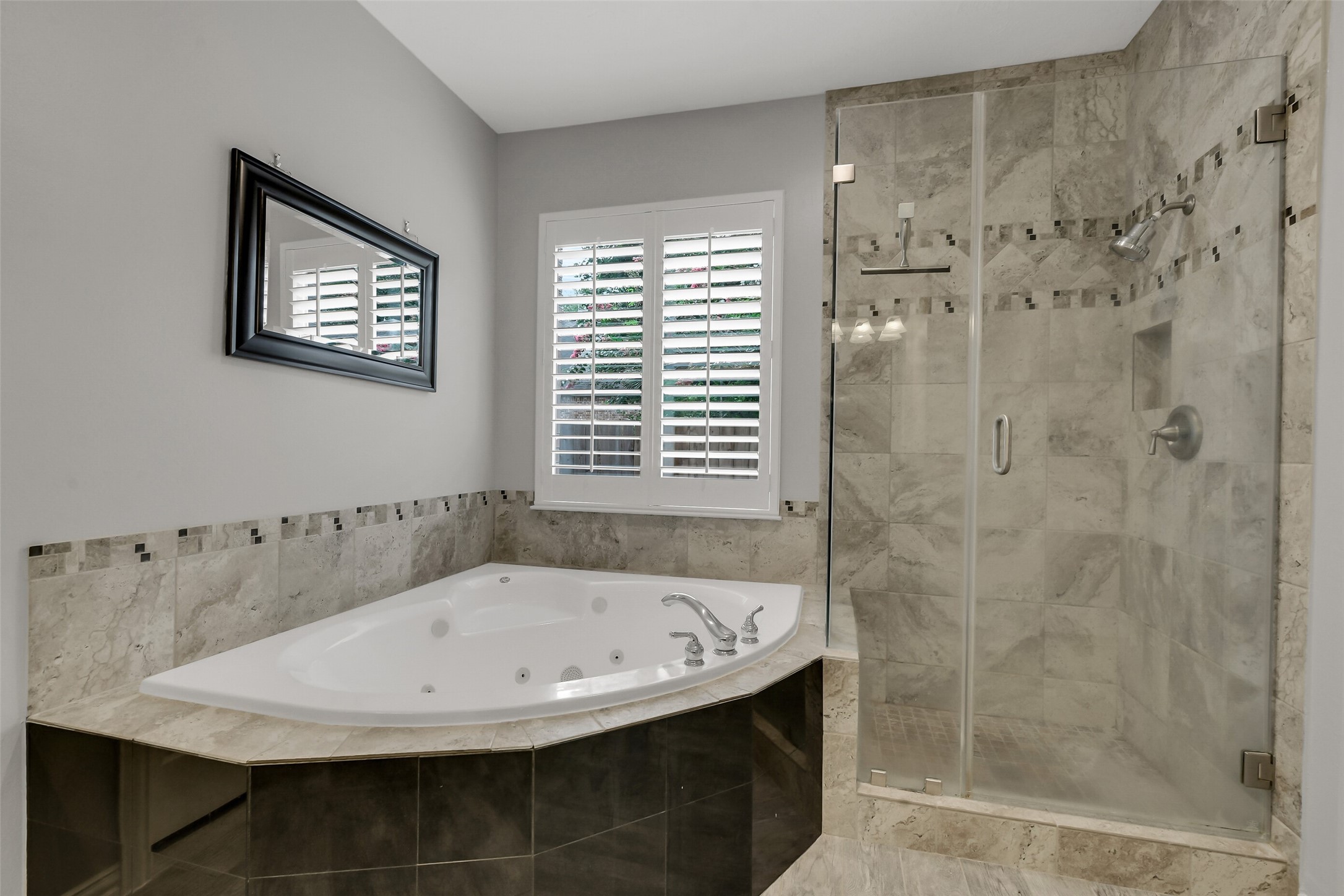 Luxurious Primary  bathroom featuring a spacious jetted tub and elegant glass-enclosed shower with stylish tile accents, complemented by a large window with plantation shutters for natural light.