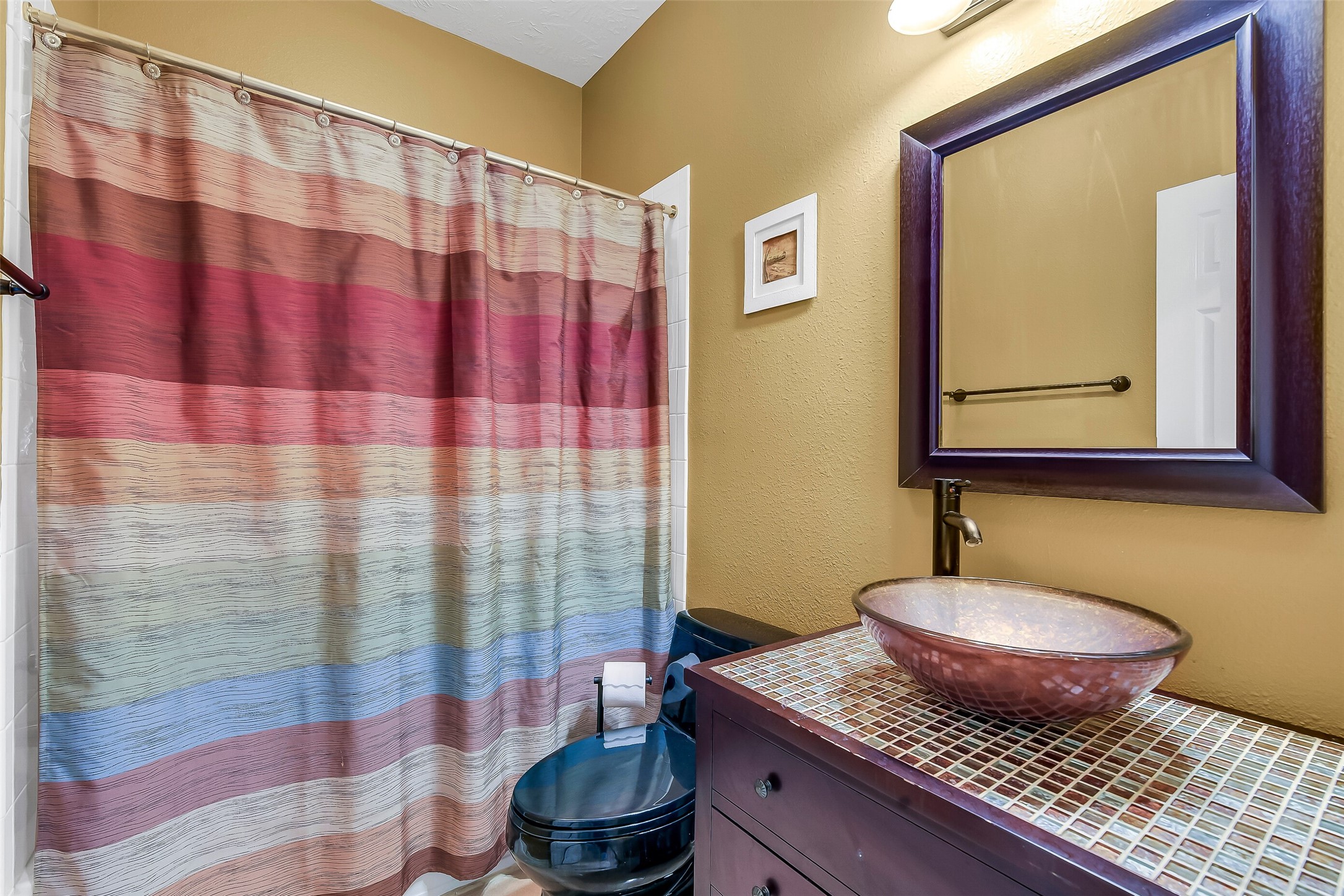 Charming bathroom located near office with a colorful striped shower curtain and a stylish vessel sink atop a tiled vanity, complemented by a warm, earthy wall color.