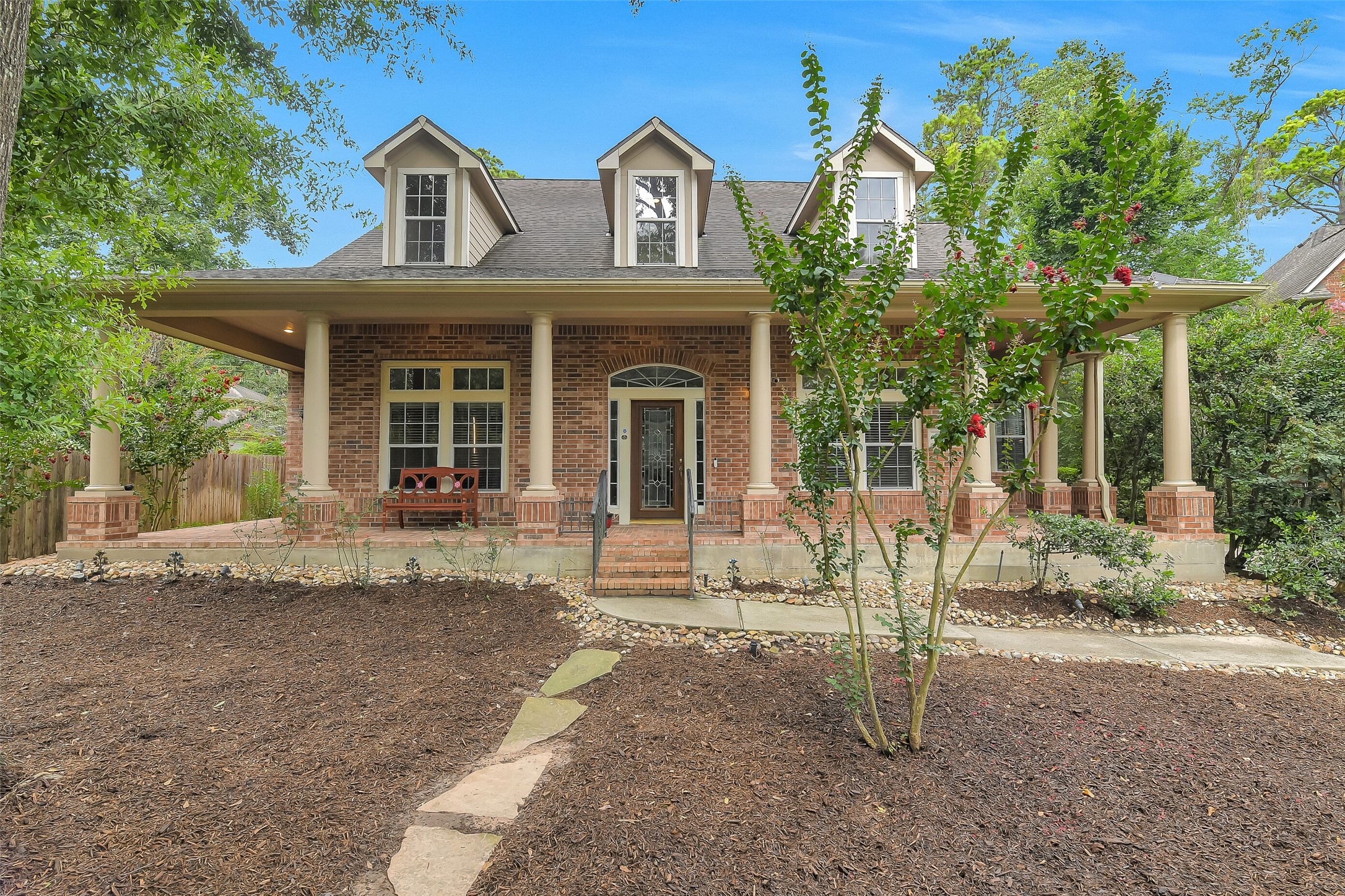 Charming brick home with a welcoming wrap-around porch, nestled amidst lush greenery and mature trees, offering a serene and inviting entrance.