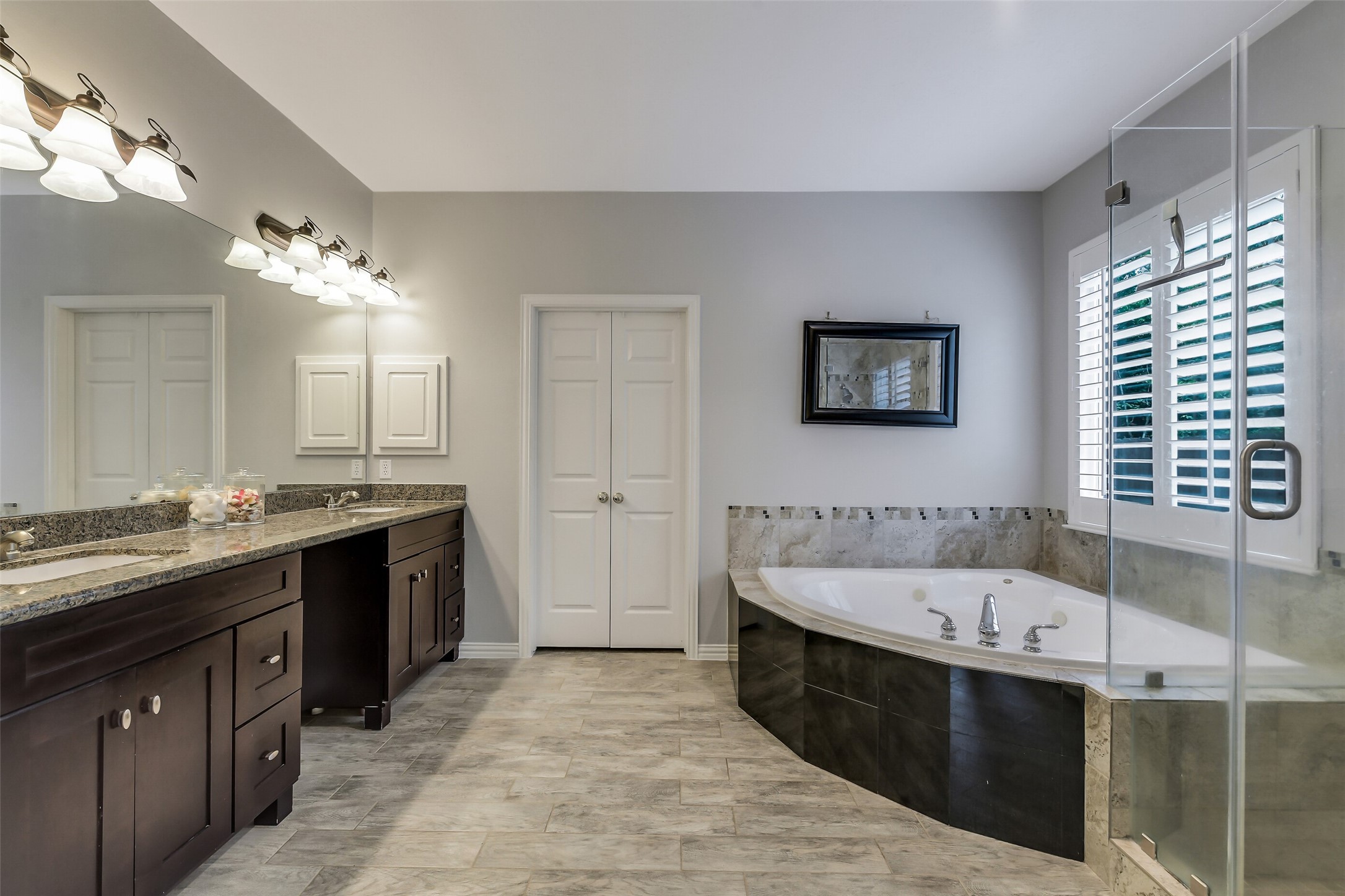 Elegant Primary  bathroom featuring a spacious dual-sink vanity with granite countertops, a luxurious corner bathtub, and a glass-enclosed shower, all enhanced by sleek tile flooring and ample natural light.