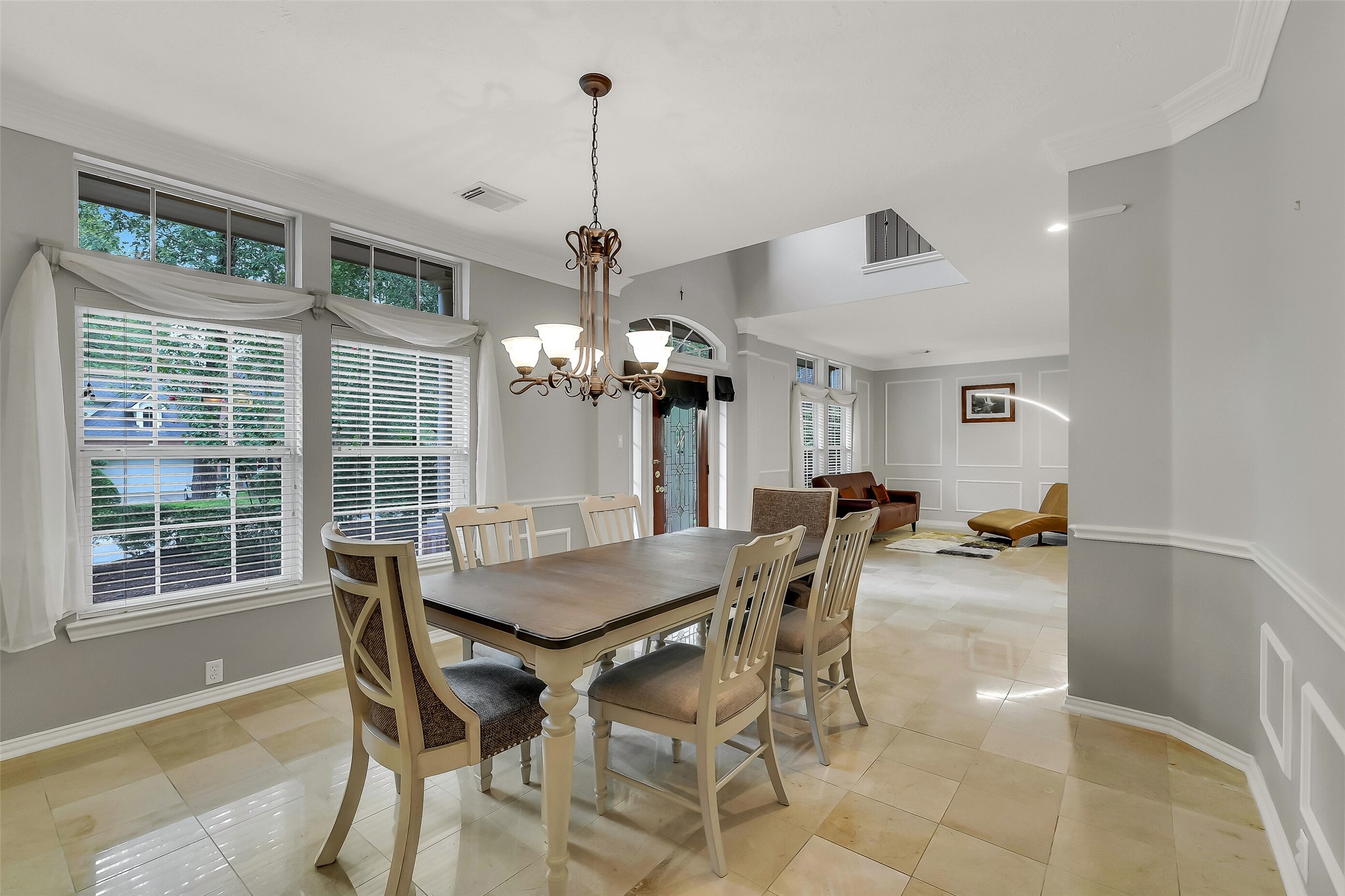 Elegant dining area with large windows, natural light, and a stylish chandelier, seamlessly connecting to a cozy living space with modern decor and marble flooring.