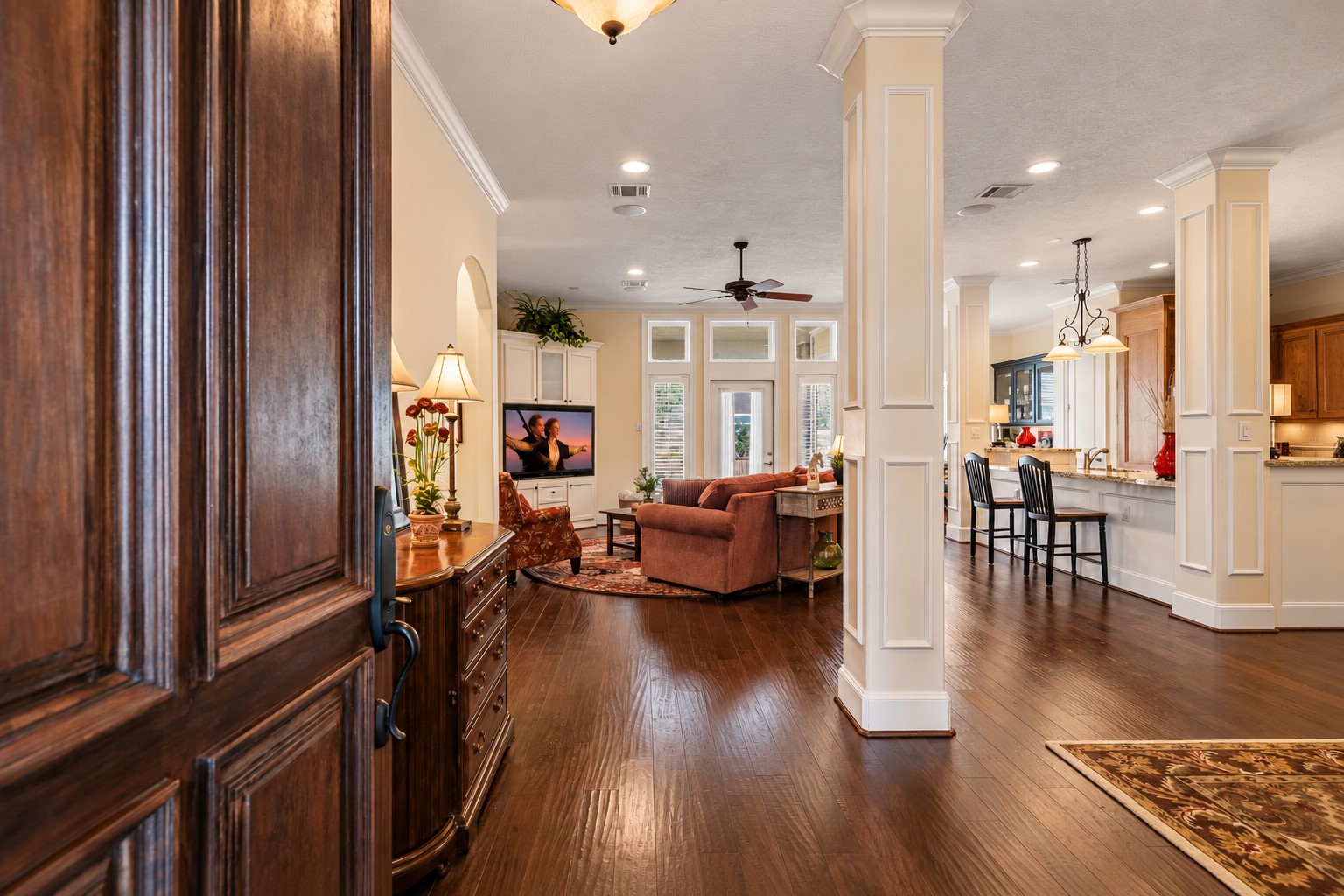 A sense of grandeur unfolds upon entry, where soaring ceilings, rich crown molding, continuous wood flooring, and stately cased columns set the tone for the home's refined design. Sunlight pours through expansive windows and dramatic transoms, illuminating the open living spaces with a warm, inviting glow. A sense of grandeur unfolds upon entry, where soaring ceilings, rich crown molding, continuous wood flooring, and stately cased columns set the tone for the home's refined design. Sunlight pours through expansive windows and dramatic transoms, illuminating the open living spaces with a warm, inviting glow.