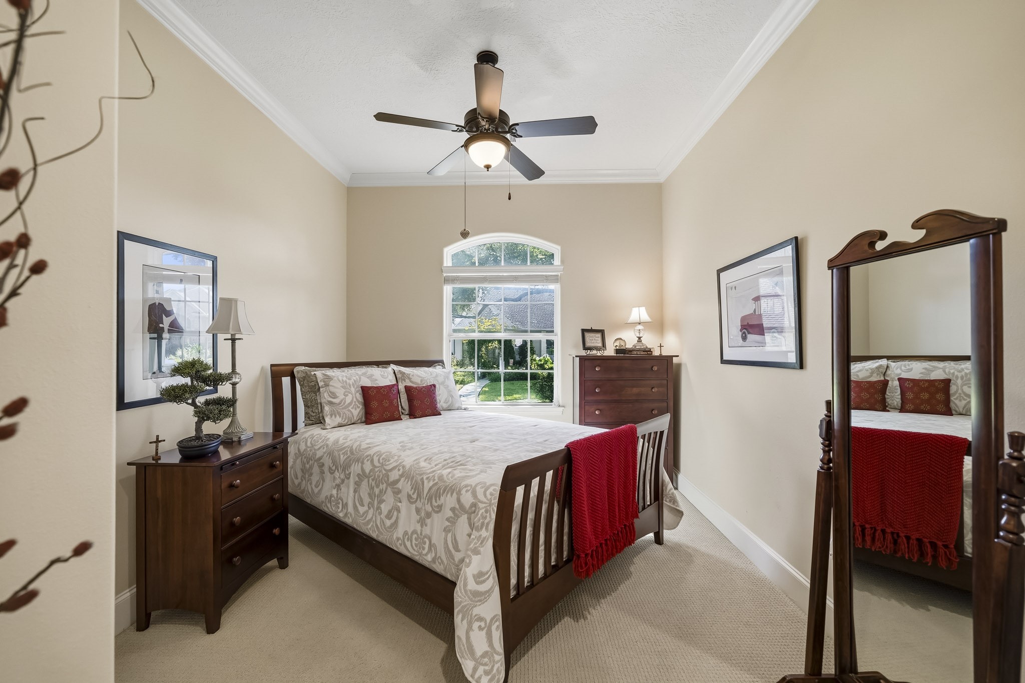 This Secondary Bedroom combines comfort and character, featuring durable carpeting, elegant crown molding, a 5-blade ceiling fan, and a spacious walk-in closet. An expansive window crowned by a graceful arched transom fills the room with natural light while framing serene views of the lush Front Yard, creating a bright and inviting retreat.