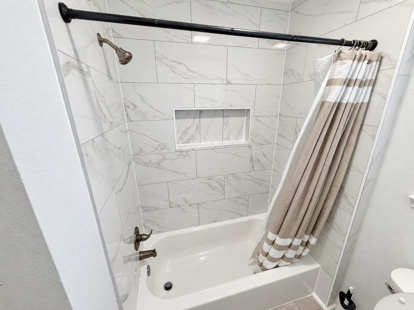 Tub/shower combination featuring marble-look tile and a recessed storage niche.