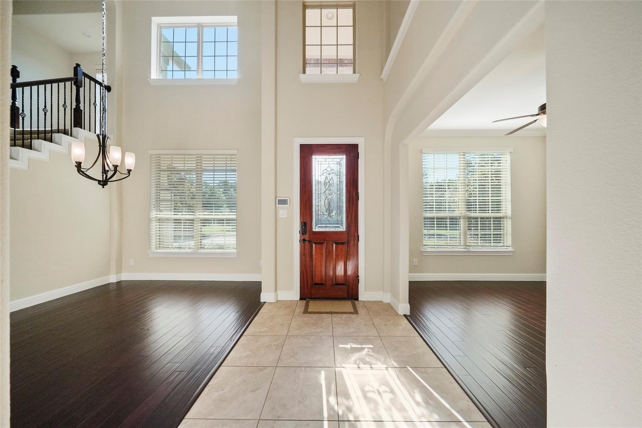 Grand two-story foyer, showing the wood front door with decorative glass, hanging chandelier, staircase with iron railing, tile entry flanked by dark wood floors, and large windows with blinds.